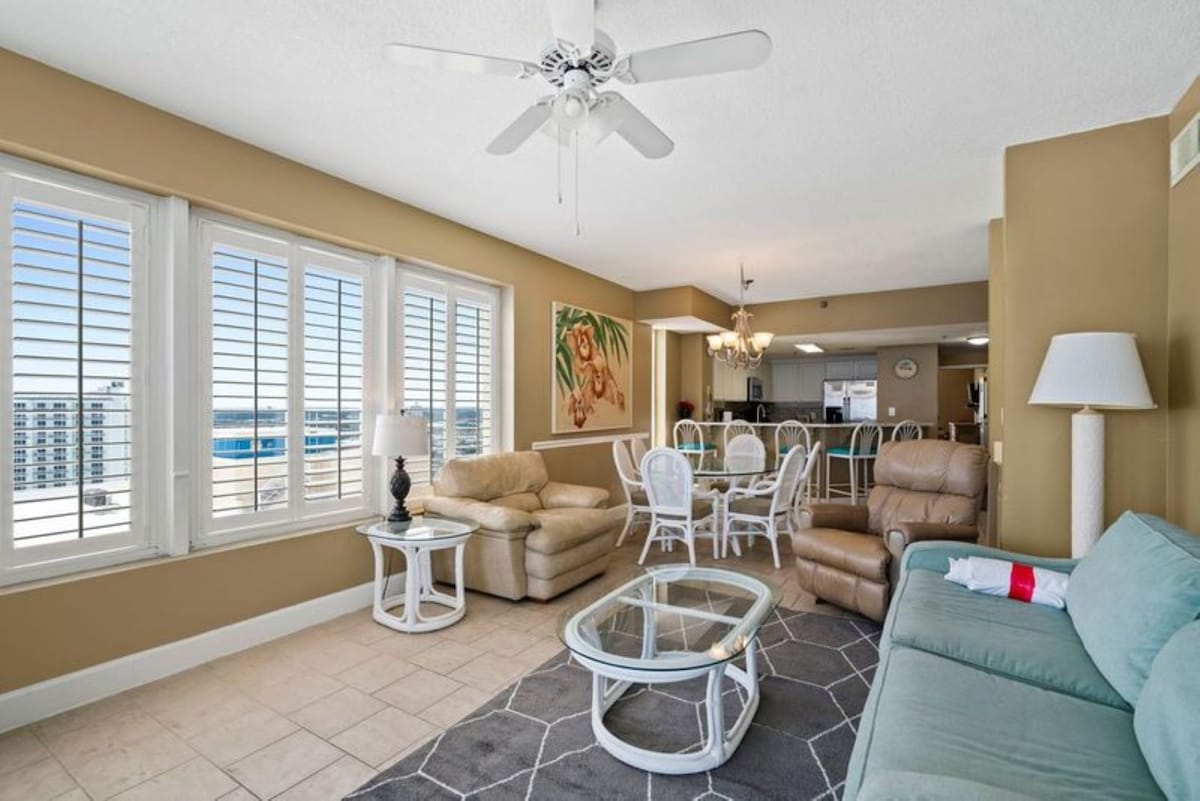An open-concept living and dining area with light tile flooring, featuring a light blue sofa, armchairs, and a dining table with white wicker chairs, all illuminated by natural light from large windows.