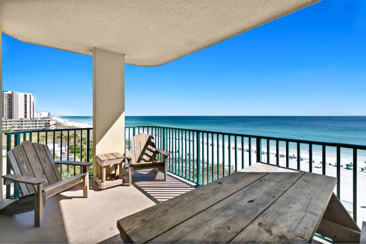 A wide balcony with stunning ocean and beach views, furnished with a wooden picnic table and two Adirondack chairs, offering ample outdoor seating.