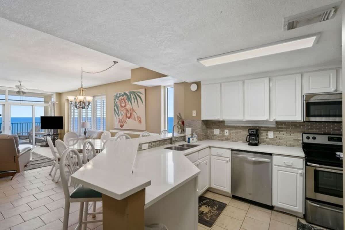 An open-concept kitchen with white cabinets, light countertops, and a mosaic tile backsplash, overlooking the dining and living areas with ocean views.