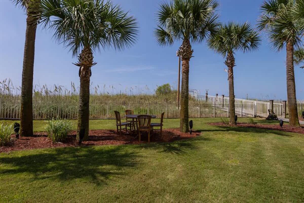 A grassy lawn area is dotted with tall palm trees, under which a wooden outdoor dining set with chairs is placed on a bed of mulch. A white fence and a boardwalk leading to the beach are visible in the background.