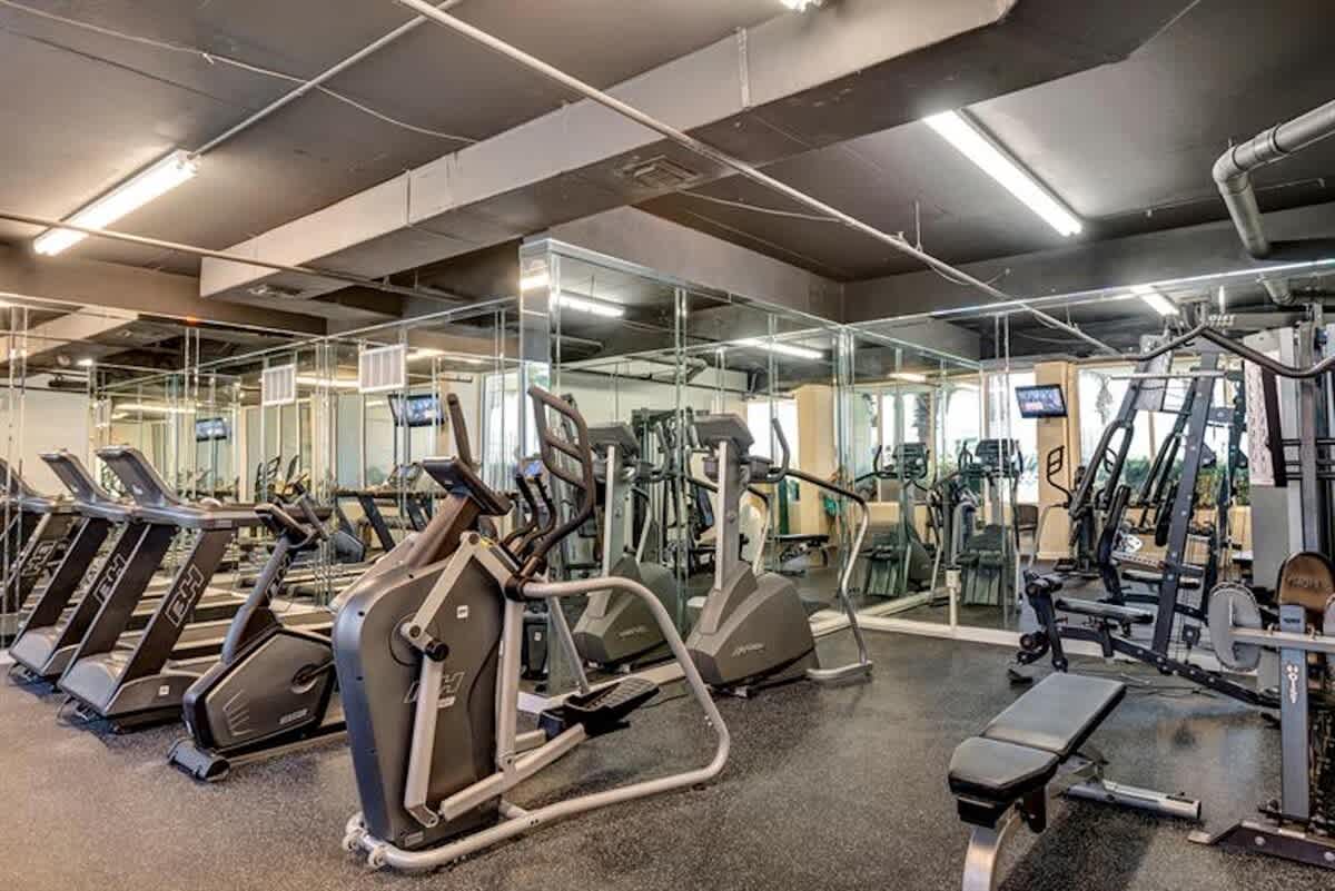 A well-equipped fitness center features a variety of cardio machines like treadmills and ellipticals, along with weightlifting equipment, all set on a black rubber floor with mirrored walls reflecting the space.