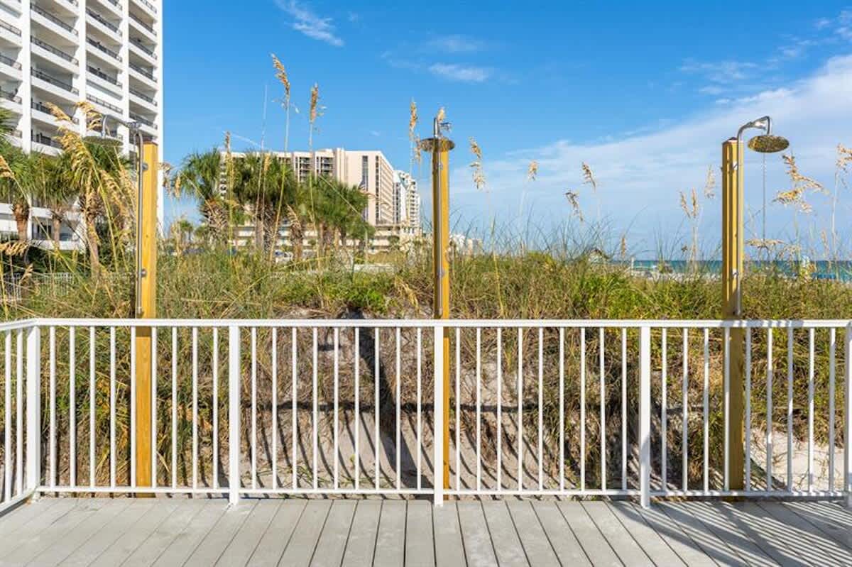 Several outdoor showers with brass showerheads are mounted on wooden posts, situated on a wooden deck overlooking dune grasses and the ocean, with resort buildings in the background.