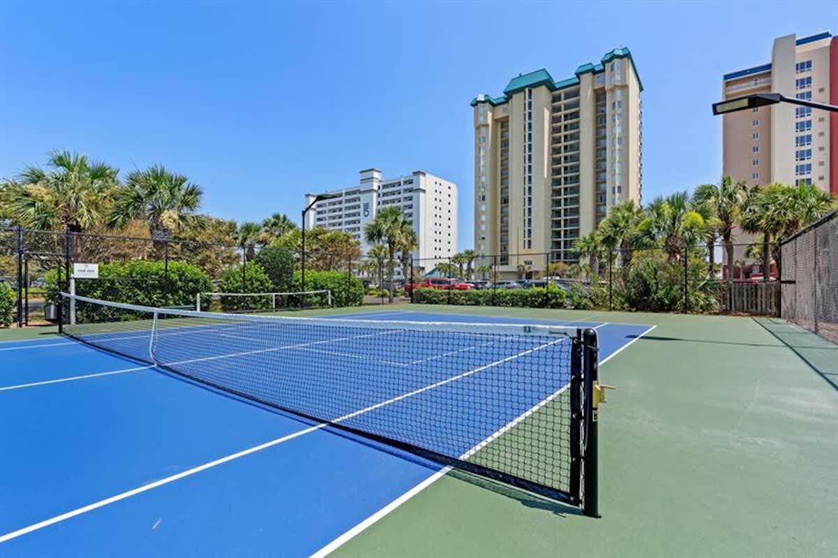 A vibrant blue and green tennis court with a net is visible, surrounded by a black chain-link fence and green landscaping, with tall resort buildings in the background under a clear blue sky.