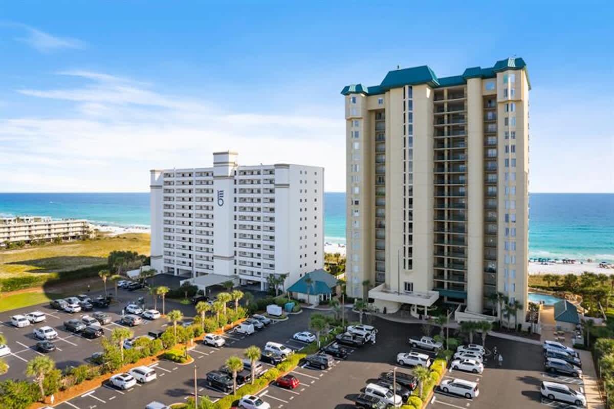An aerial view showcases two large beachfront condominium buildings with extensive parking lots filled with cars, situated directly on a white sand beach next to turquoise ocean waters. The surrounding area includes other buildings and lush greenery.