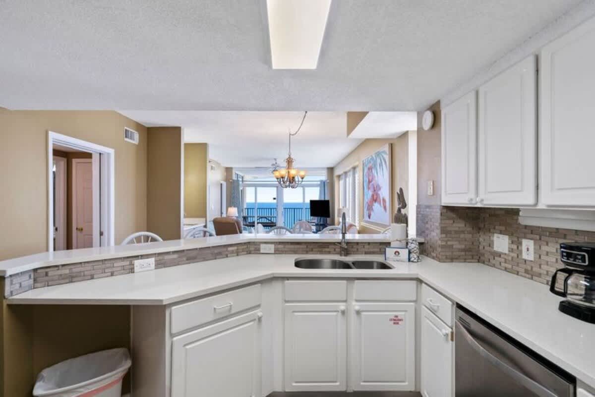 A close-up view of the kitchen's L-shaped counter with white cabinets and light countertops, featuring a double sink and a mosaic tile backsplash, with the living area visible in the background.