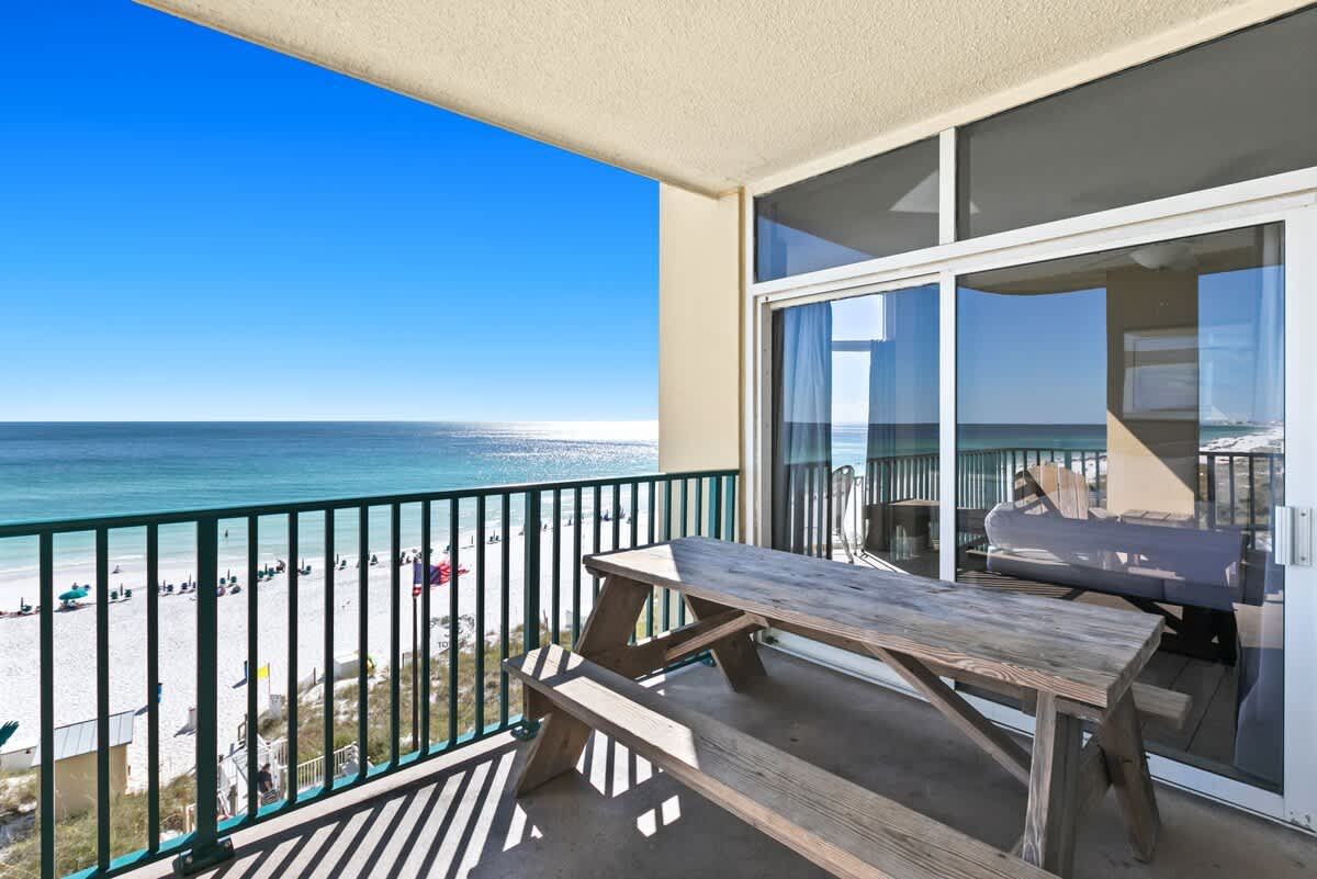 A spacious balcony overlooking a white sand beach and the turquoise ocean, featuring a rustic wooden picnic table and sliding glass doors leading indoors.