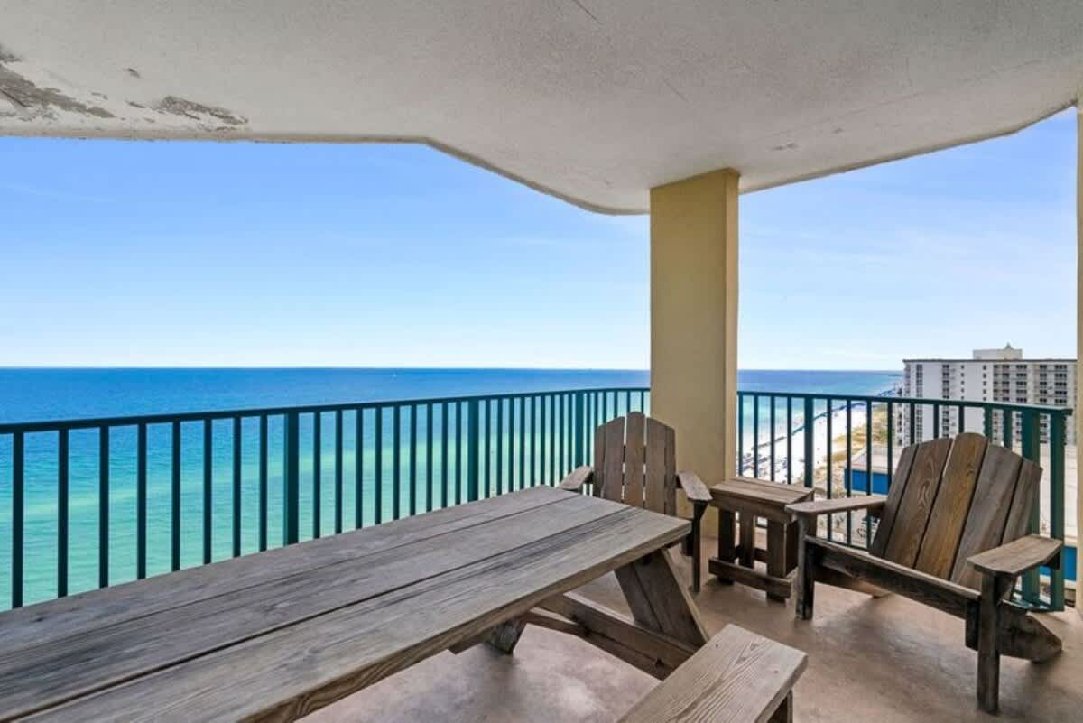Spacious balcony featuring a rustic wooden picnic table and two Adirondack chairs, offering expansive panoramic views of the turquoise ocean and distant sandy beach under a bright blue sky.
