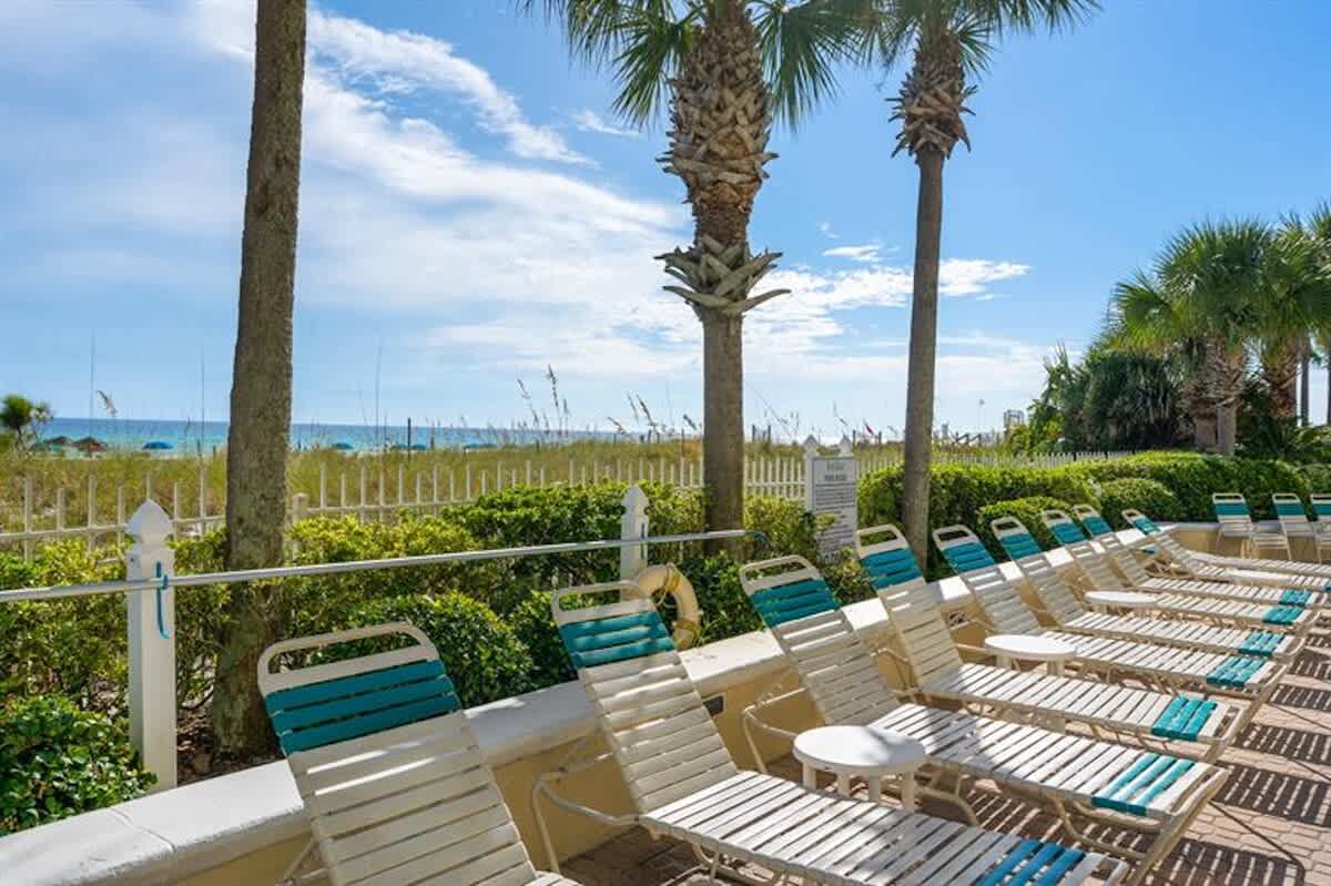 A row of white and teal lounge chairs are neatly arranged on a paved deck, offering direct views of the sandy beach and ocean beyond a white fence and dune grasses, framed by tall palm trees.