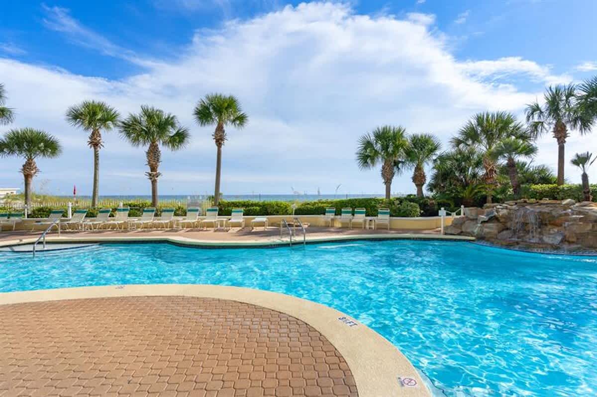 A large, curved outdoor swimming pool with a shallow entry area, surrounded by a brick paver deck and numerous lounge chairs, with palm trees and the ocean visible in the background under a blue sky.