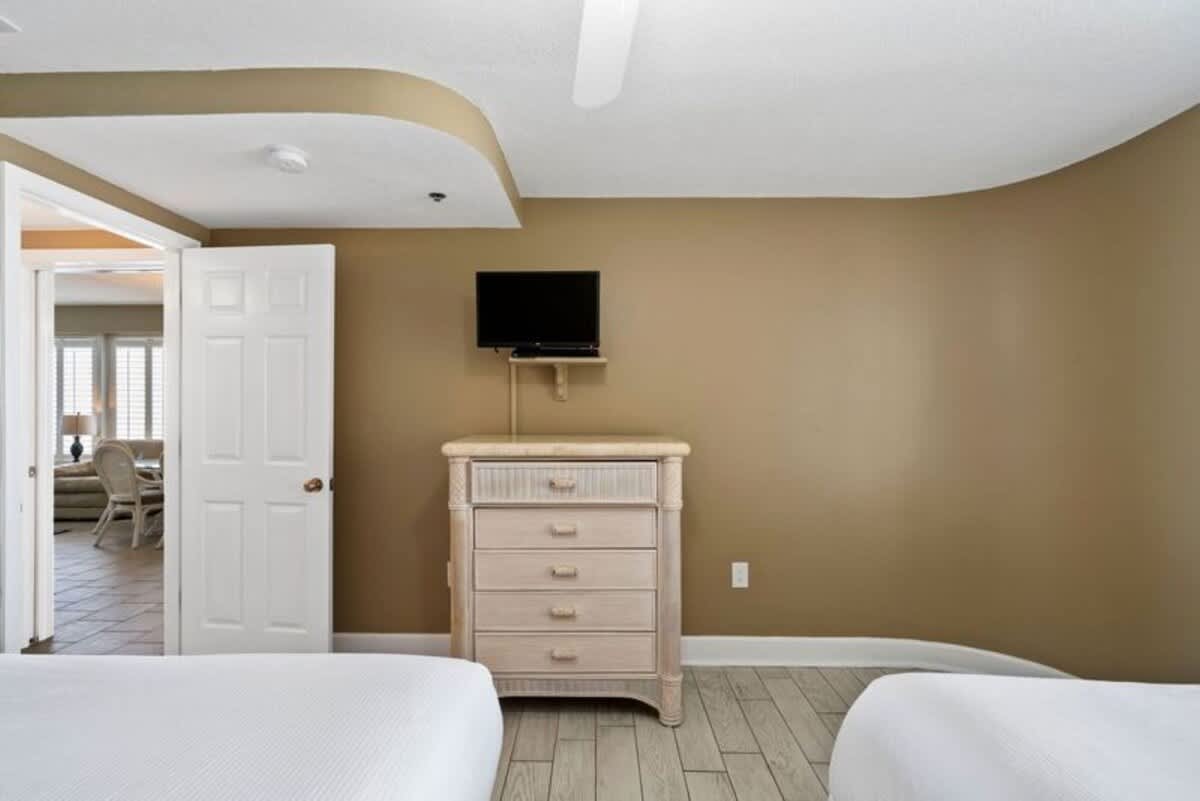 A bedroom wall with a light-colored, wicker-style dresser and a small wall-mounted television above it. Light wood-look flooring and a white door leading to another room are visible.