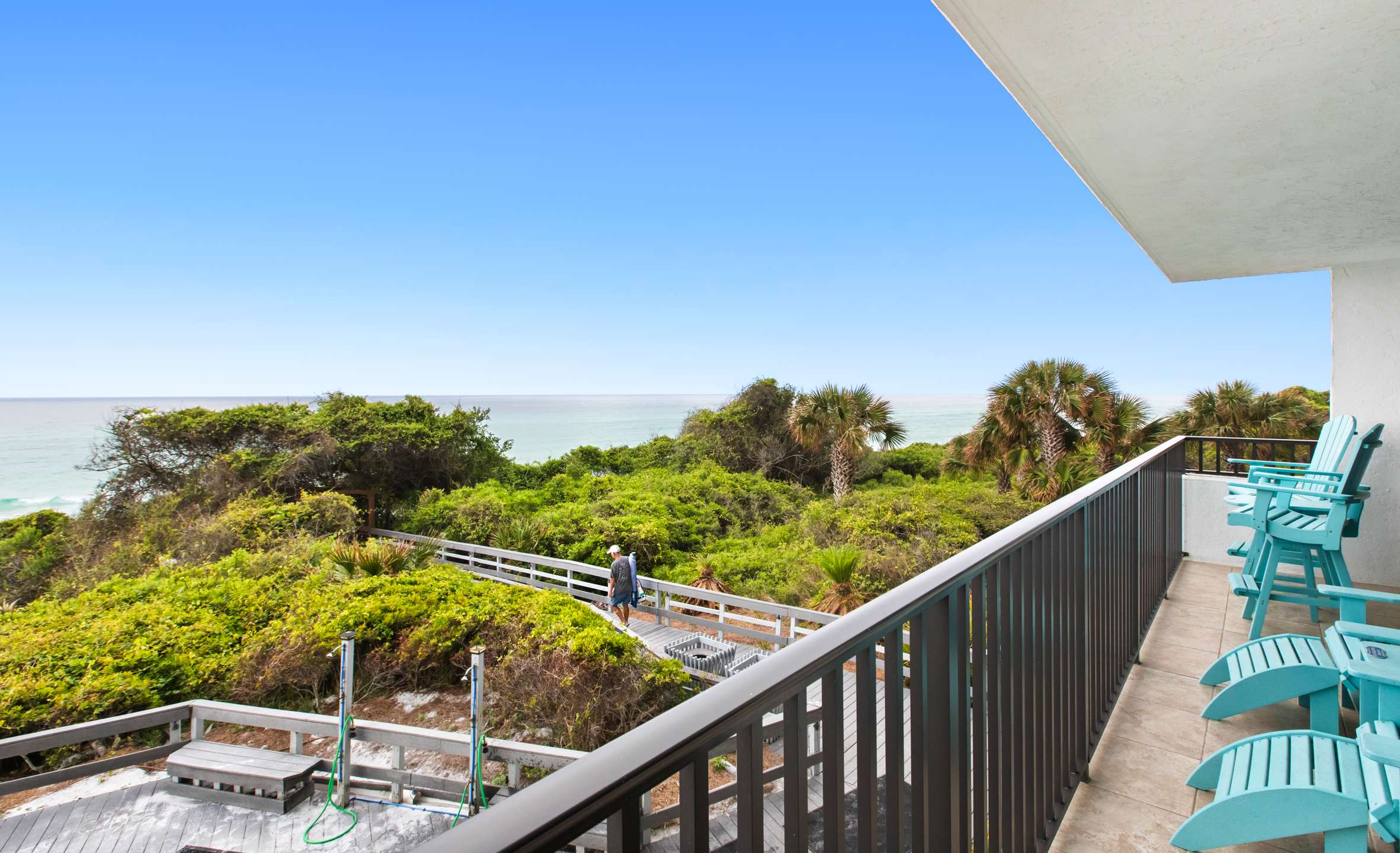 Private balcony with a dark metal railing and tiled floor, featuring two light blue Adirondack chairs. It overlooks a lush green landscape, a wooden boardwalk, and the sparkling ocean under a clear blue sky.