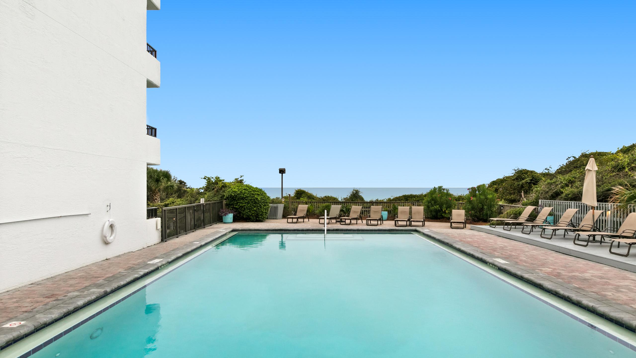 Rectangular outdoor swimming pool with a light blue interior, surrounded by a brick paver deck and several lounge chairs. The ocean is visible in the background, beyond a fence and some greenery.