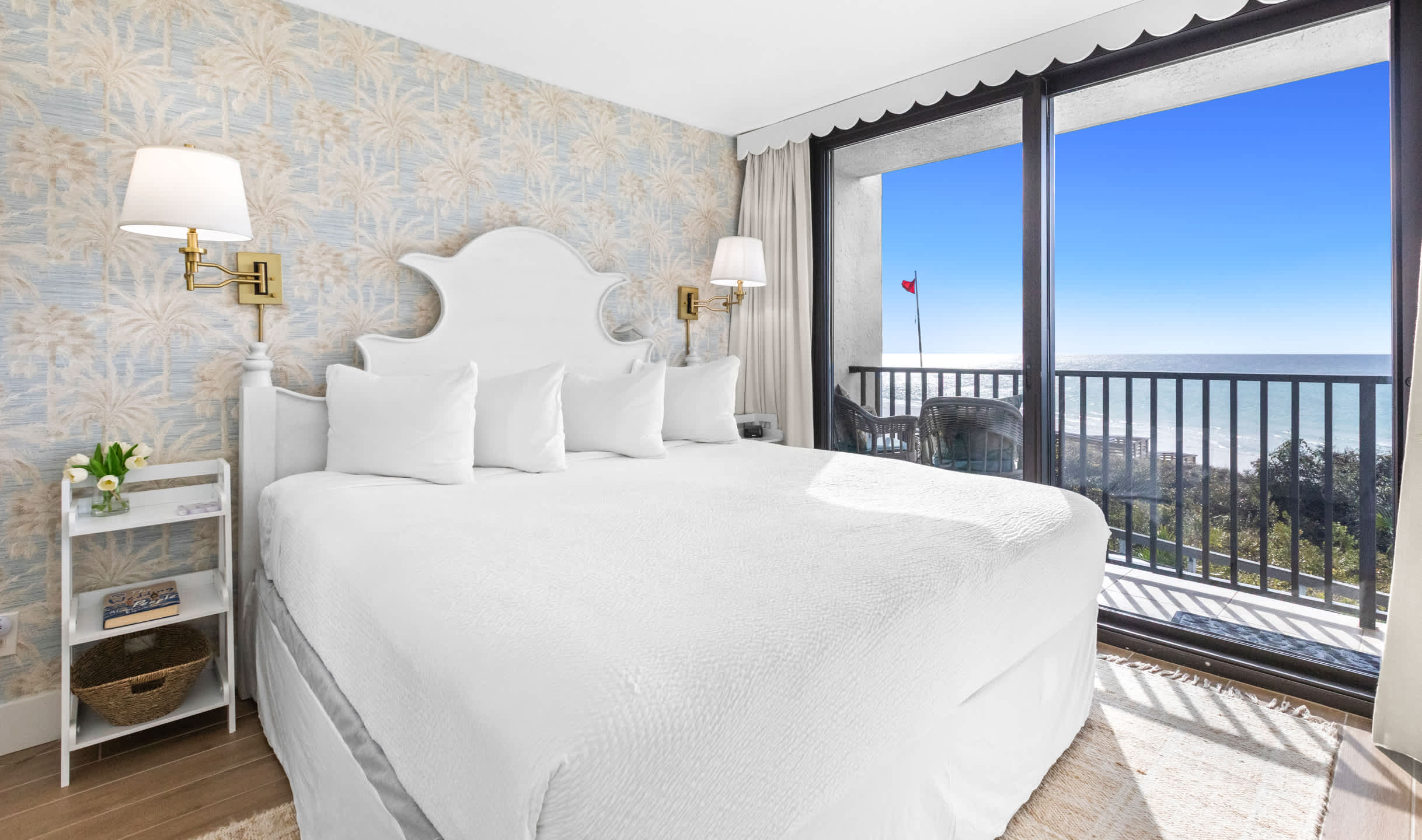 Master bedroom featuring a white ornate headboard on a king bed with white bedding, palm tree patterned wallpaper, and direct balcony access with stunning ocean views.