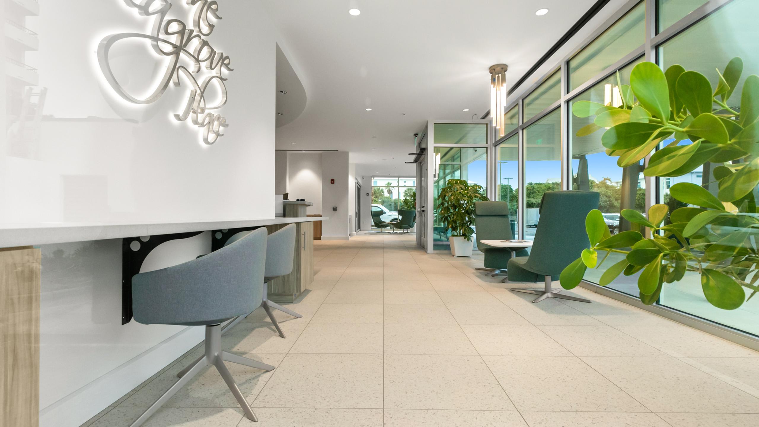 A contemporary lobby space with light tiled flooring, featuring a long counter with bar stools, modern green armchairs, and large windows looking out to the exterior.