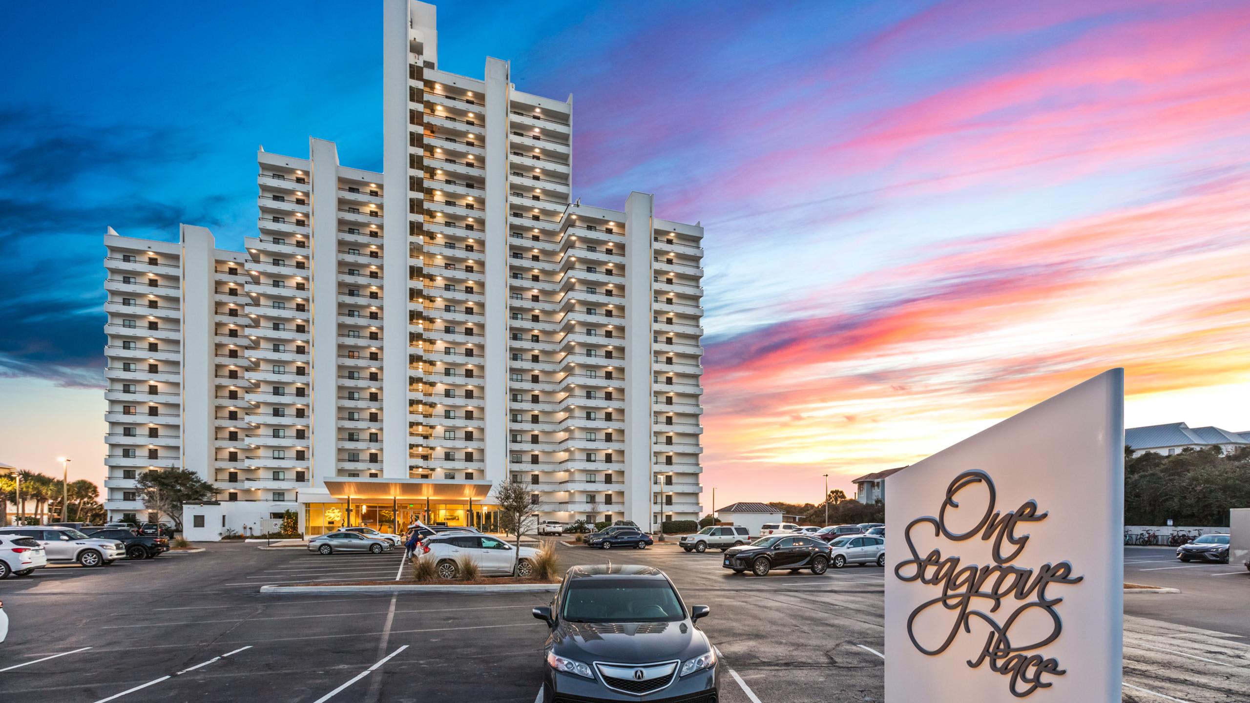A tall, multi-story white condominium building stands prominently against a vibrant sunset sky, with a parking lot in the foreground and a "One Seagrove Place" sign.