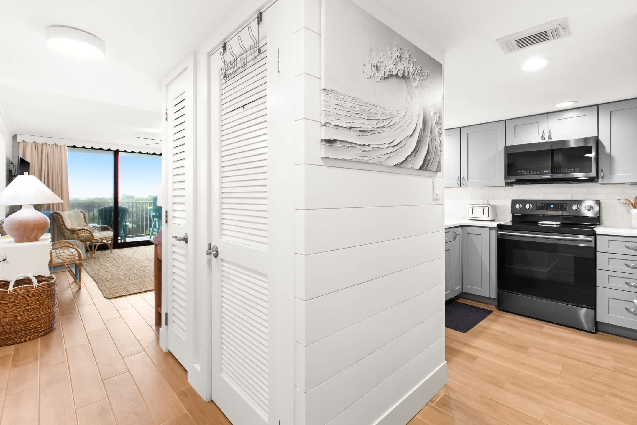Bright hallway with light wood-look flooring and white shiplap walls, featuring a large wave art piece and leading into the kitchen and living area with ocean views.