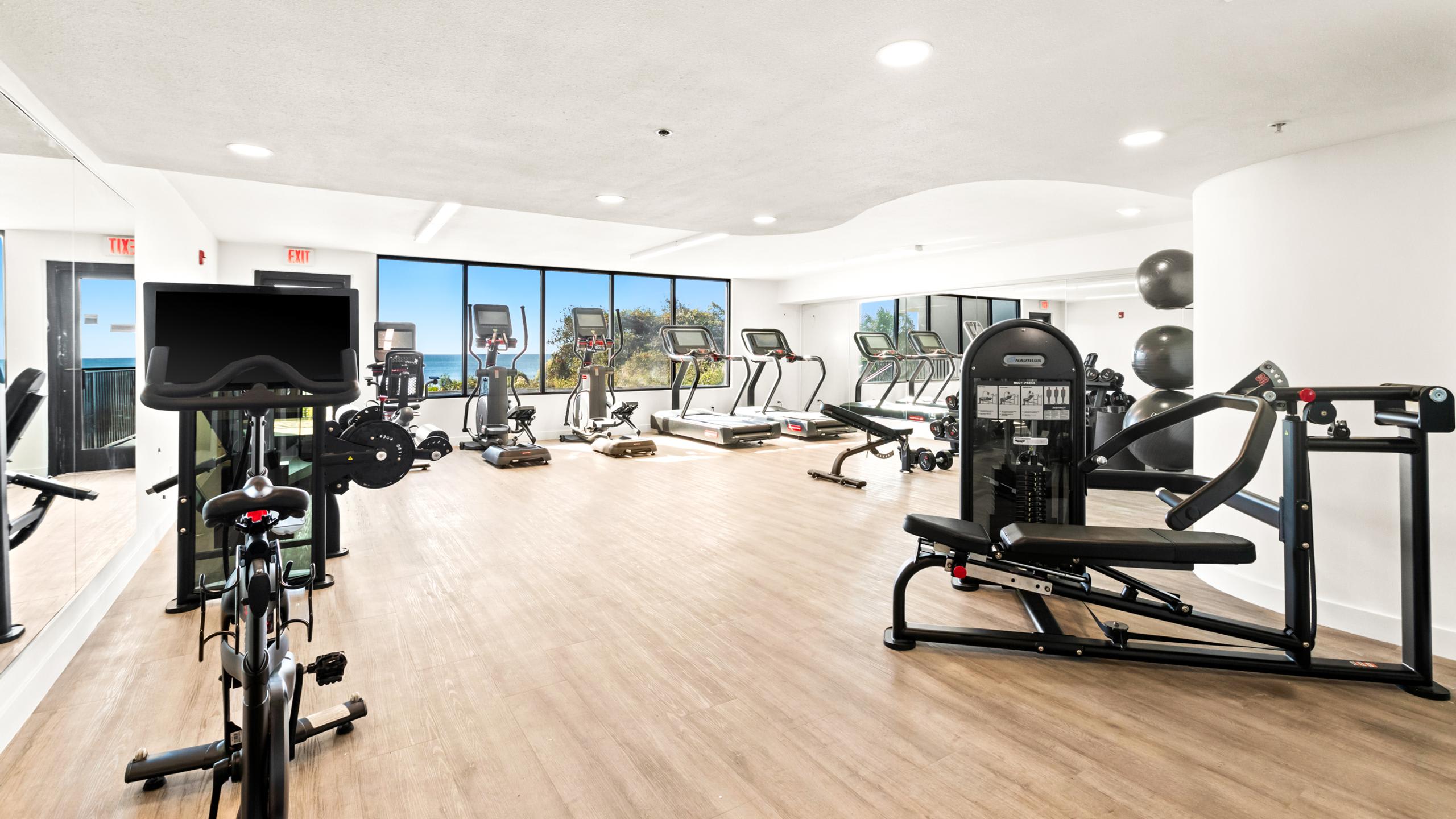 A modern fitness center with light wood-look flooring and white walls, equipped with weight machines, exercise bikes, and treadmills, all facing large windows with an ocean view.