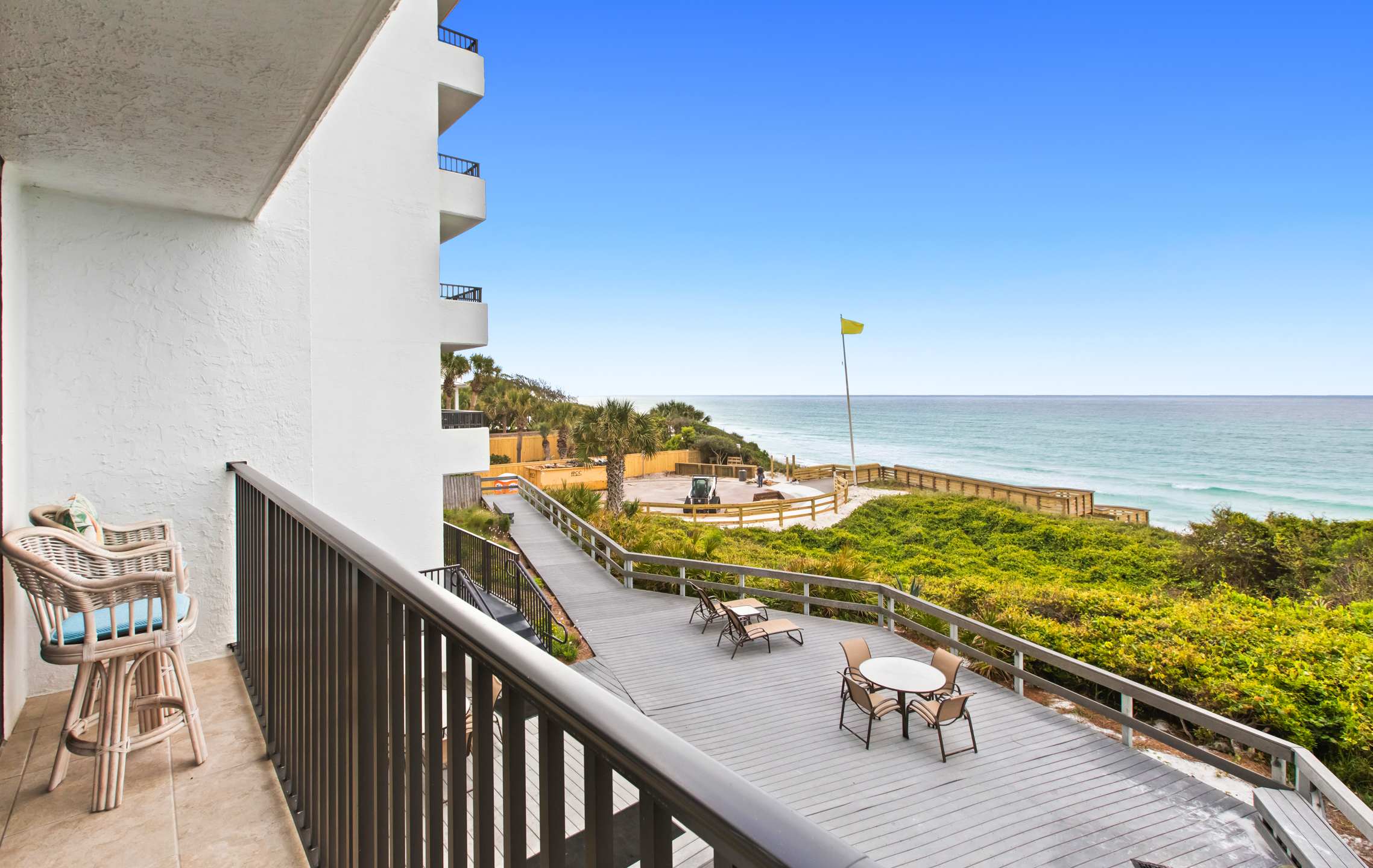 Balcony with a dark metal railing and a wicker bar stool, providing a view of a large wooden deck with outdoor seating, a boardwalk, and the ocean beyond. A white building facade is visible to the left.