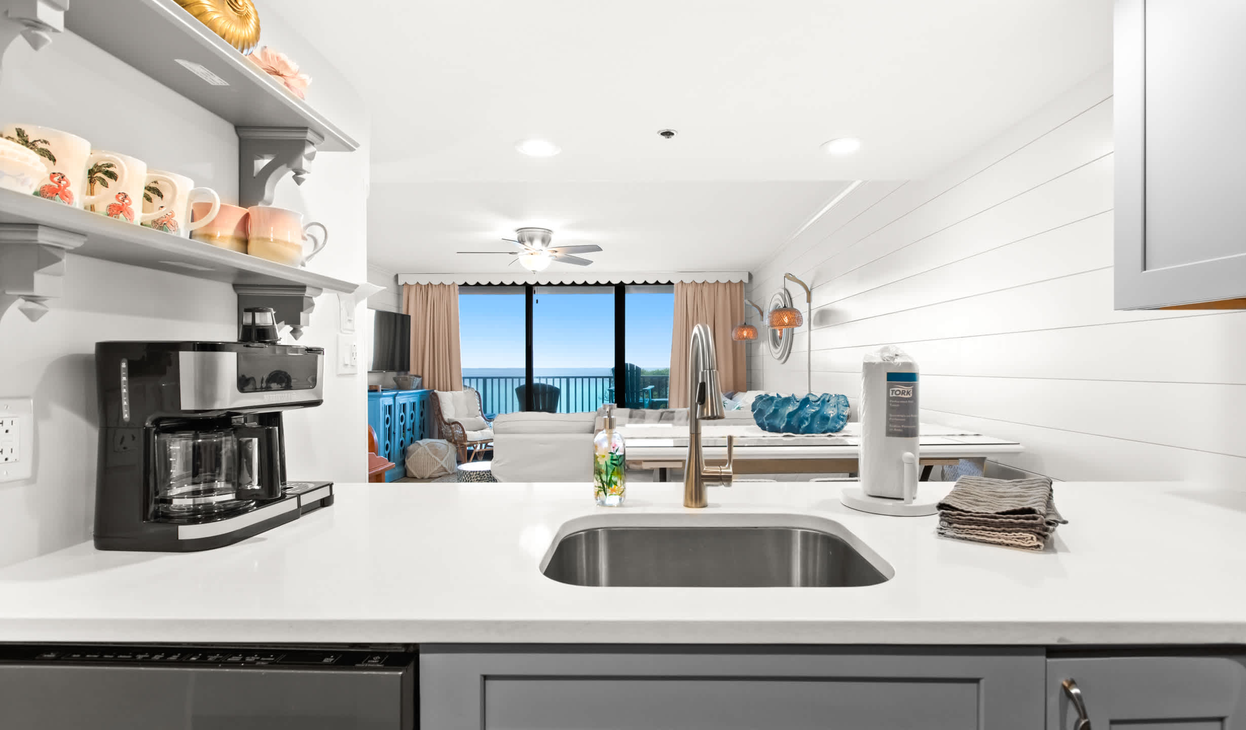 Modern kitchen with white countertops, a stainless steel sink, and a black coffee maker, offering a clear view into the living area and ocean beyond through large windows.