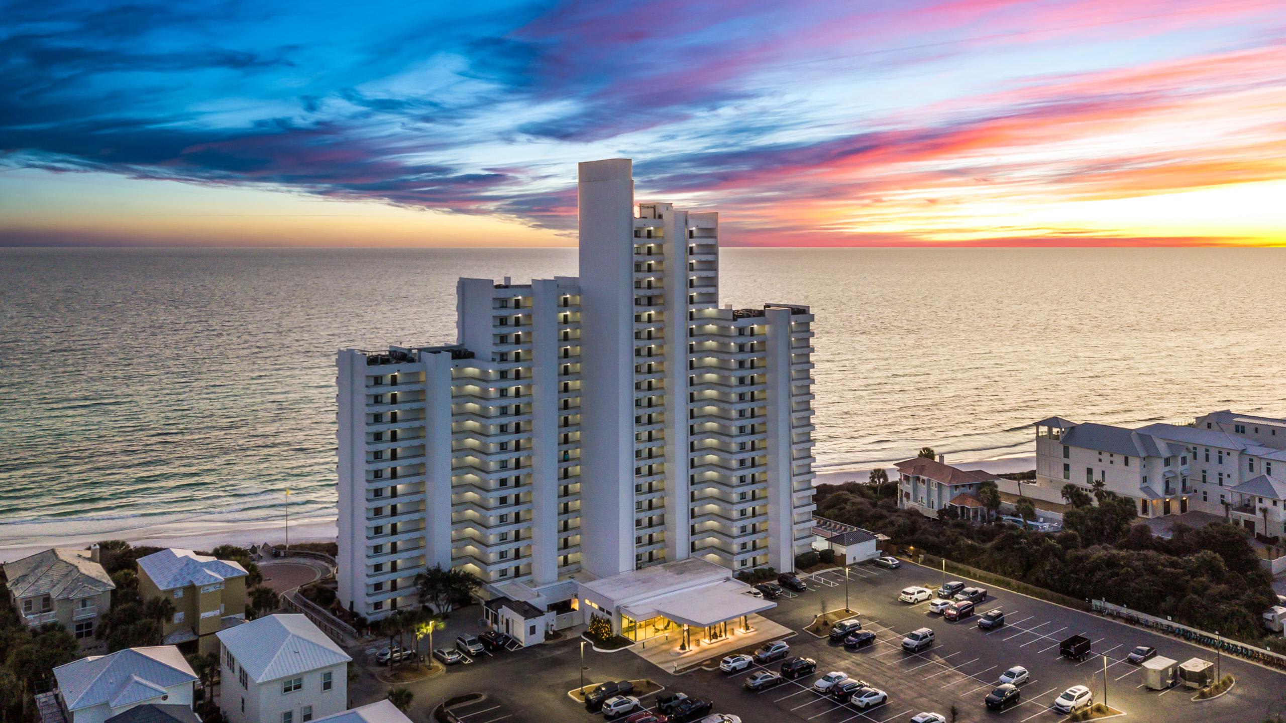 An aerial view of a tall, white high-rise condominium building situated directly on a sandy beach, with the vast ocean stretching to the horizon under a dramatic sunset sky. A parking lot with cars is visible at the base, alongside smaller residential structures.