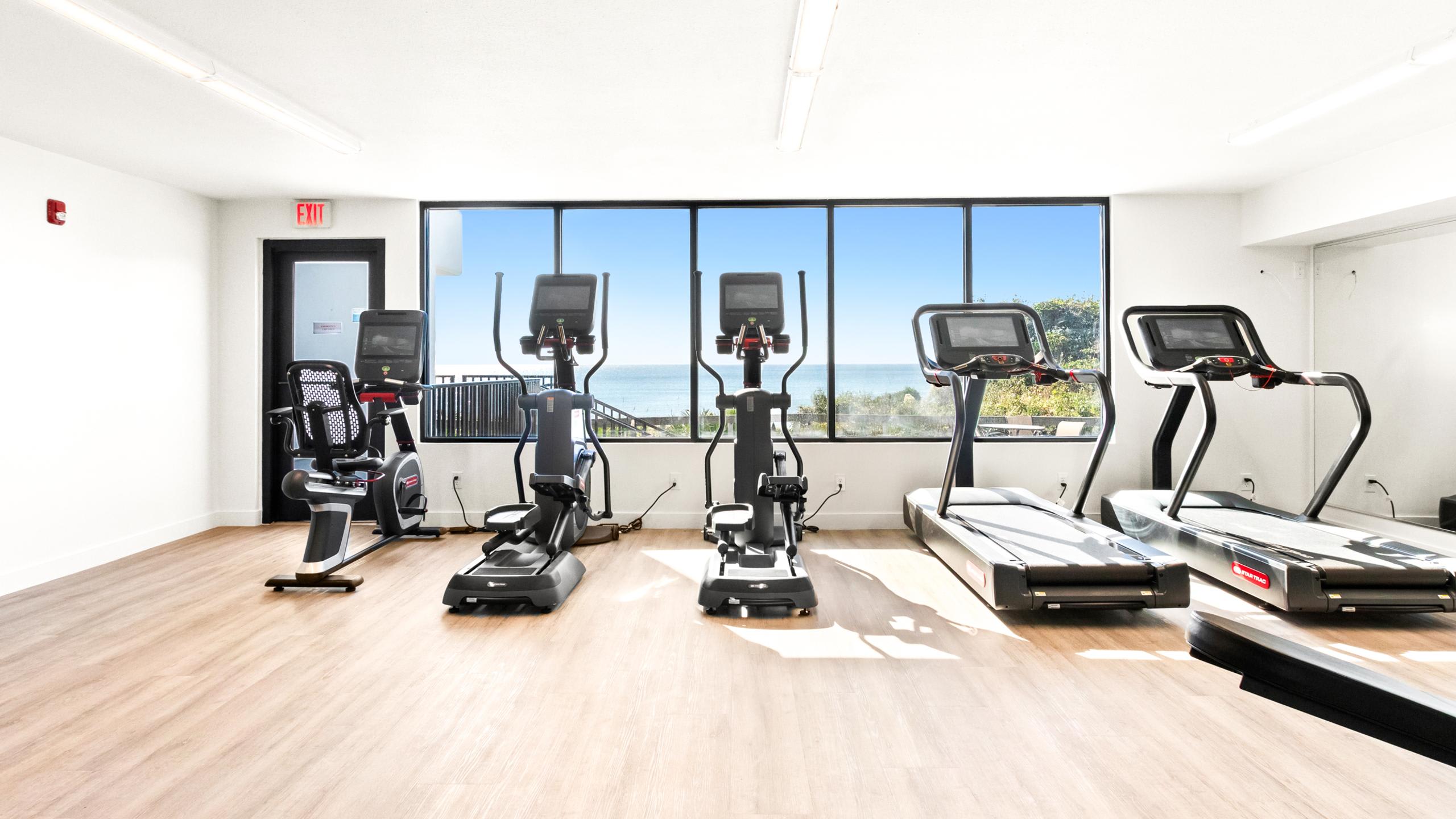 A bright fitness room features several cardio machines, including ellipticals and treadmills, positioned to offer panoramic ocean views through large windows.