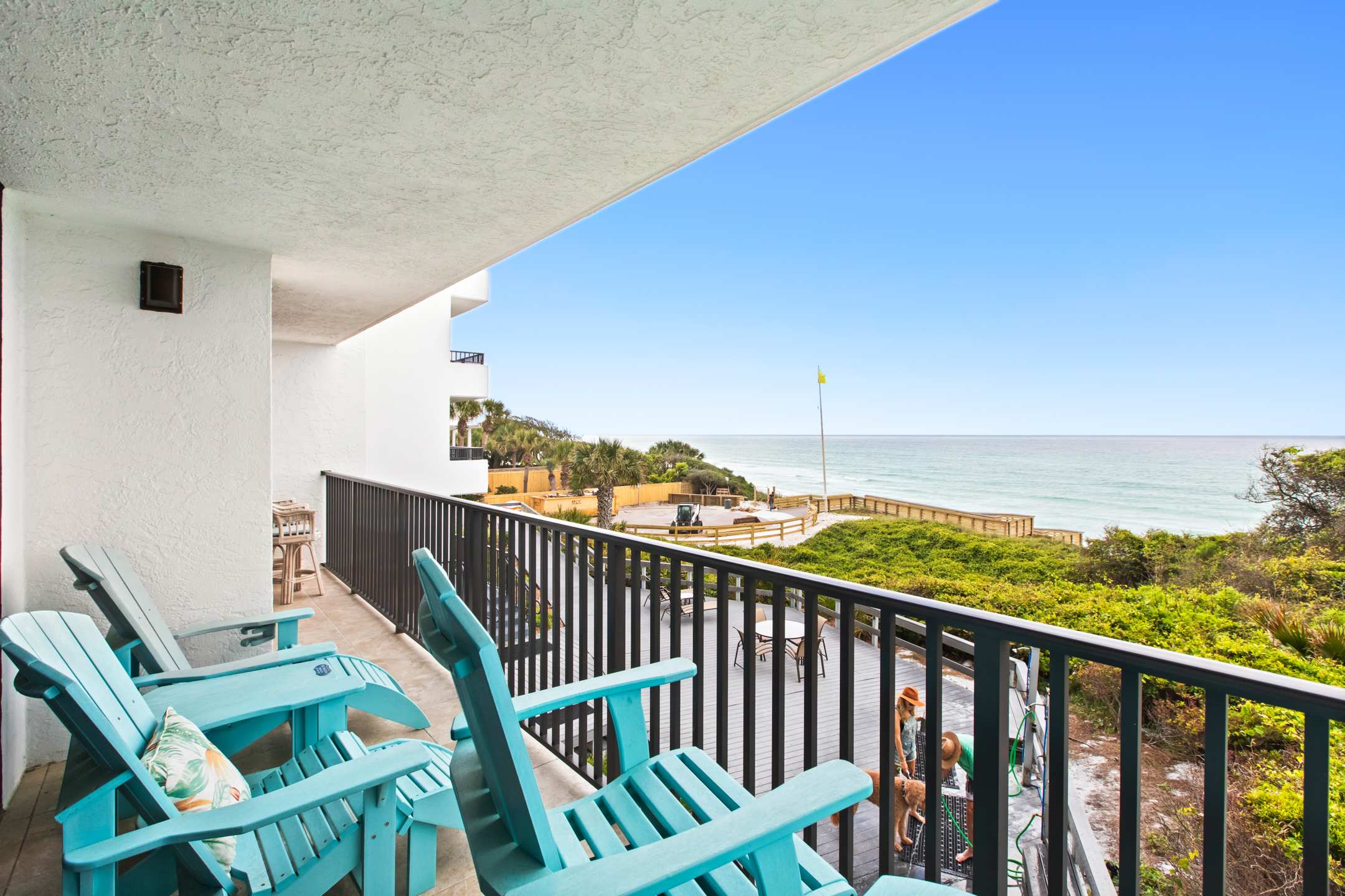 Balcony with a dark metal railing and light blue Adirondack chairs, overlooking a large wooden deck with outdoor seating, a boardwalk, and the ocean. The white building facade extends to the left, with other balconies visible.