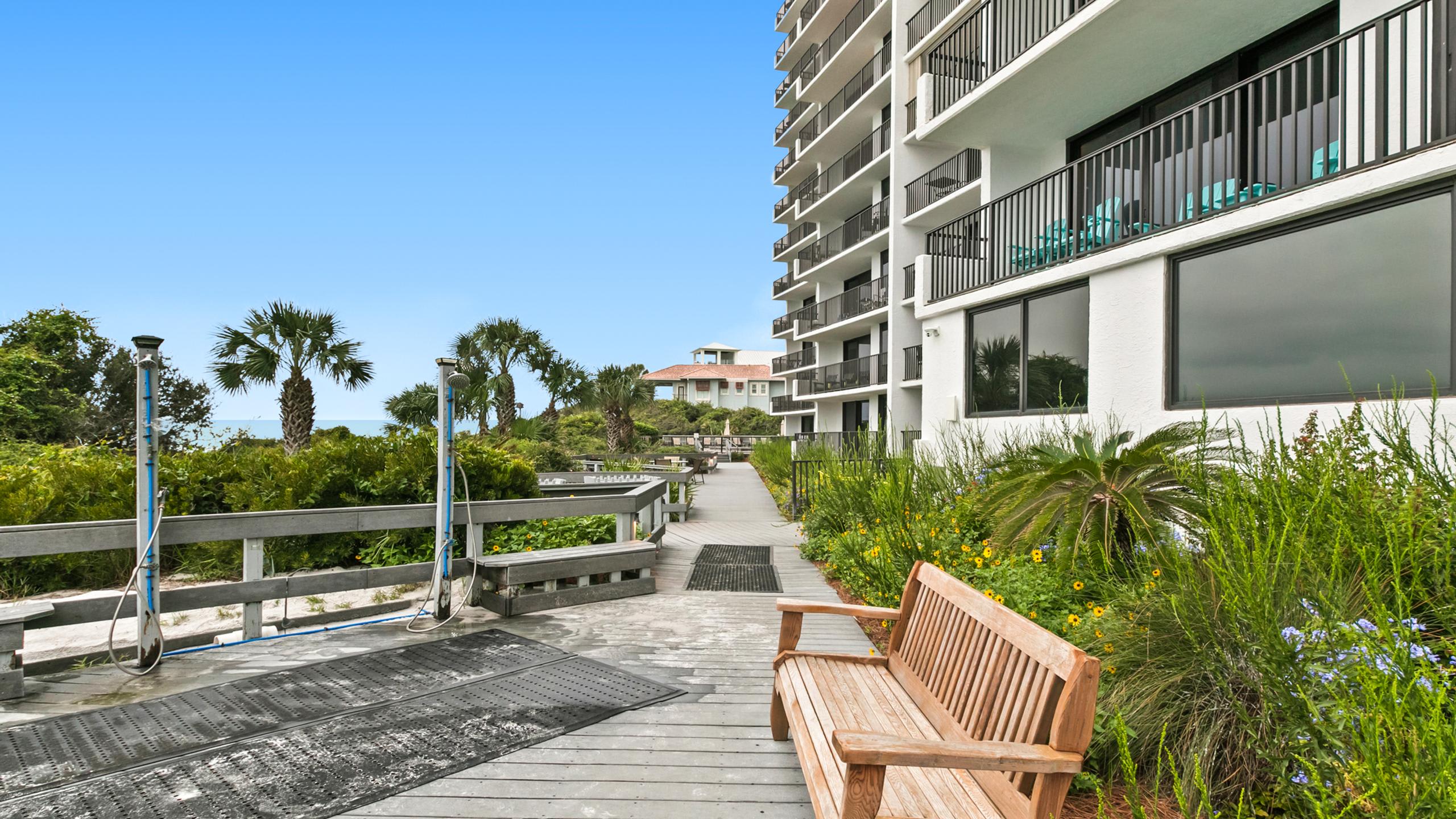 A wooden boardwalk with outdoor showers and benches, leading towards the ocean, flanked by lush greenery and a tall white condominium building with balconies.