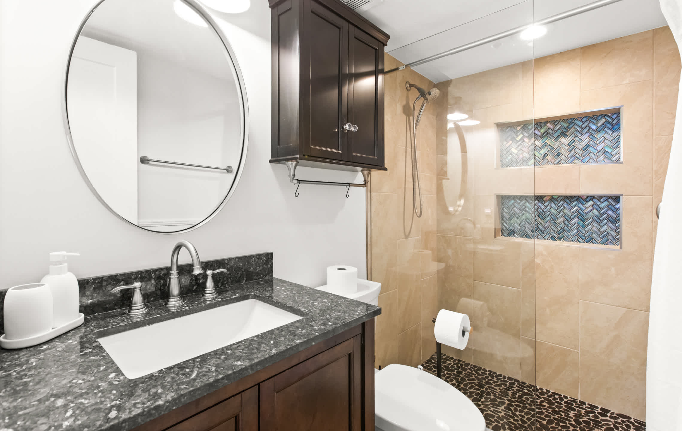 Modern bathroom featuring a dark wood vanity with a speckled granite countertop, a white rectangular sink, and a large round mirror. The walk-in shower has beige tiled walls, a glass enclosure, mosaic tile niches, and a dark pebble floor.
