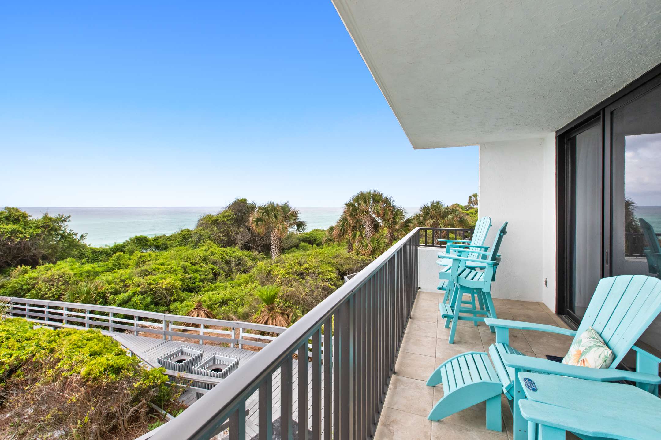 Private balcony with a dark metal railing and light blue Adirondack chairs, offering a clear view of the ocean, a wooden boardwalk, and dense green vegetation. The white exterior wall of the building is visible to the left.
