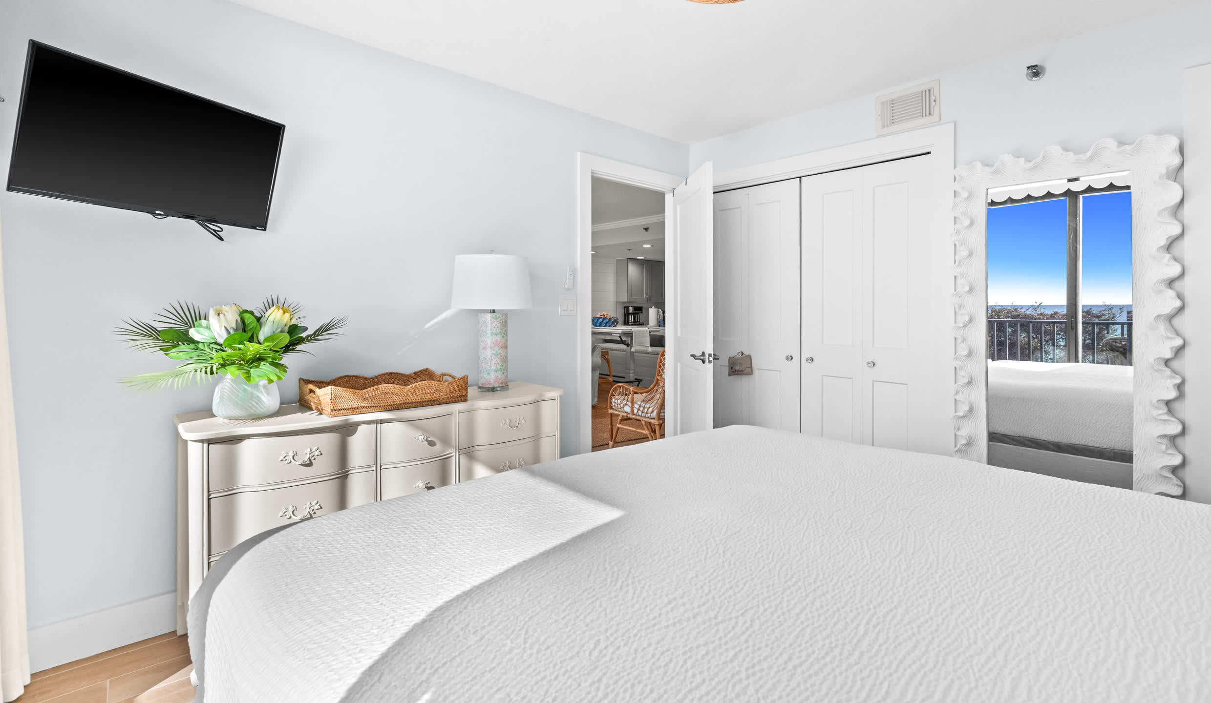 View of the master bedroom from the foot of the bed, showing a white dresser with a lamp and flowers, a wall-mounted TV, and an open doorway leading to the main living area.