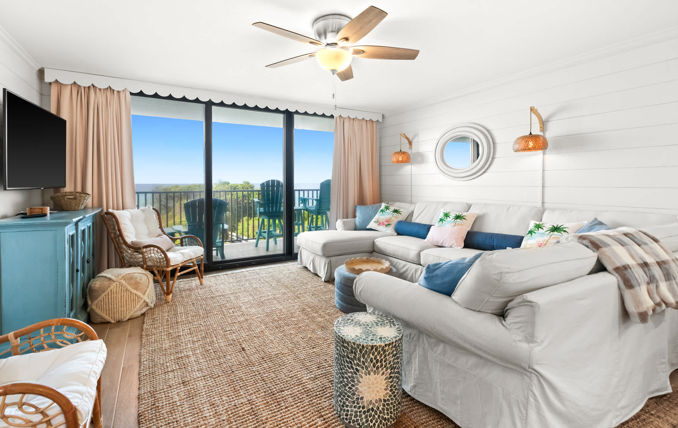 Spacious living room with a white sectional sofa, a natural fiber rug, and a view of the ocean from the balcony, complemented by shiplap walls and a round decorative mirror.