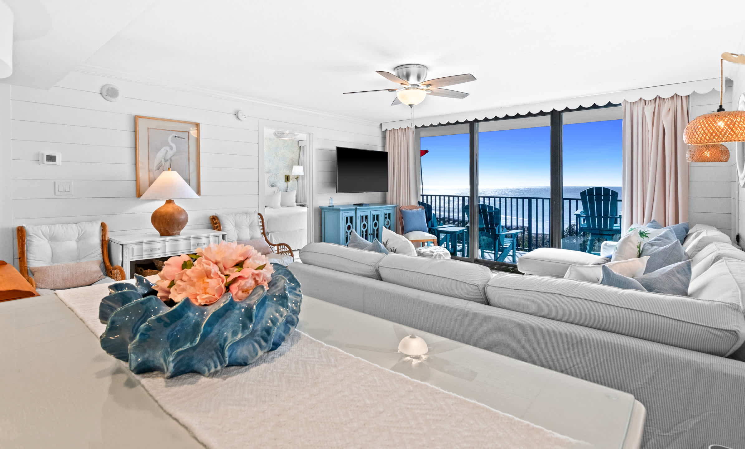 Expansive living room with a large white sectional sofa facing a balcony with ocean views, featuring shiplap walls, a light blue console, and a decorative ceramic bowl on a counter in the foreground.
