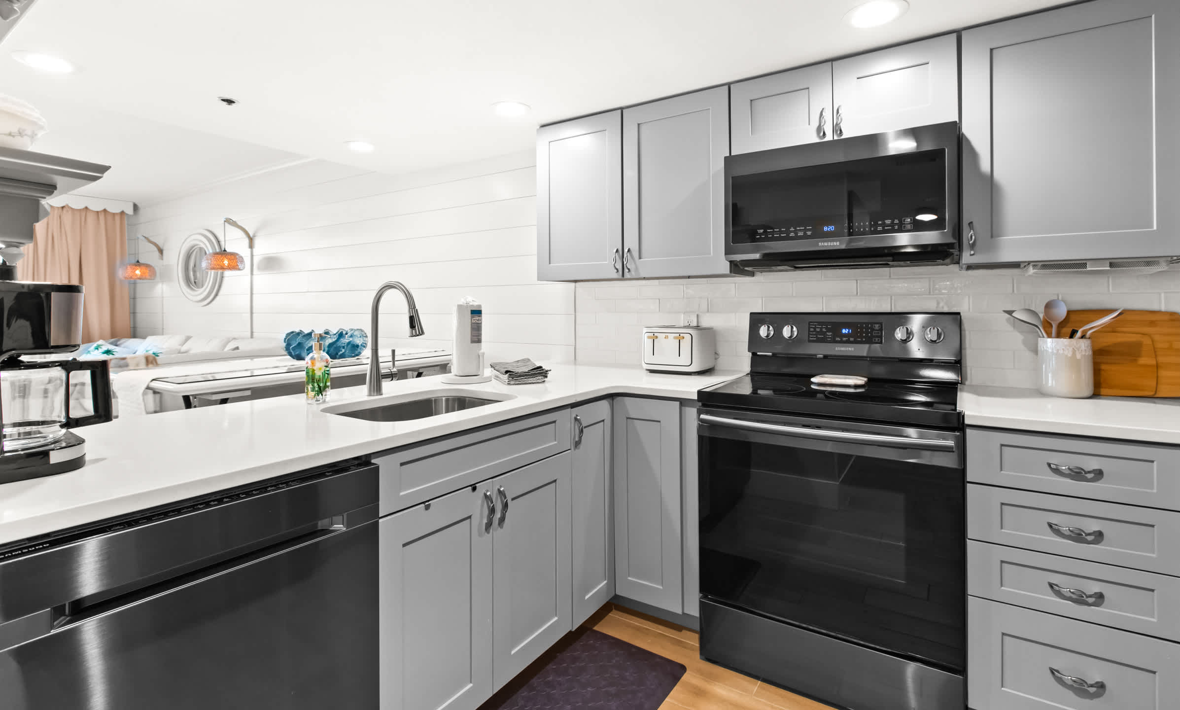 Wide shot of a contemporary kitchen with light gray cabinets, white countertops, a stainless steel sink, and black appliances, opening up to a bright living space.