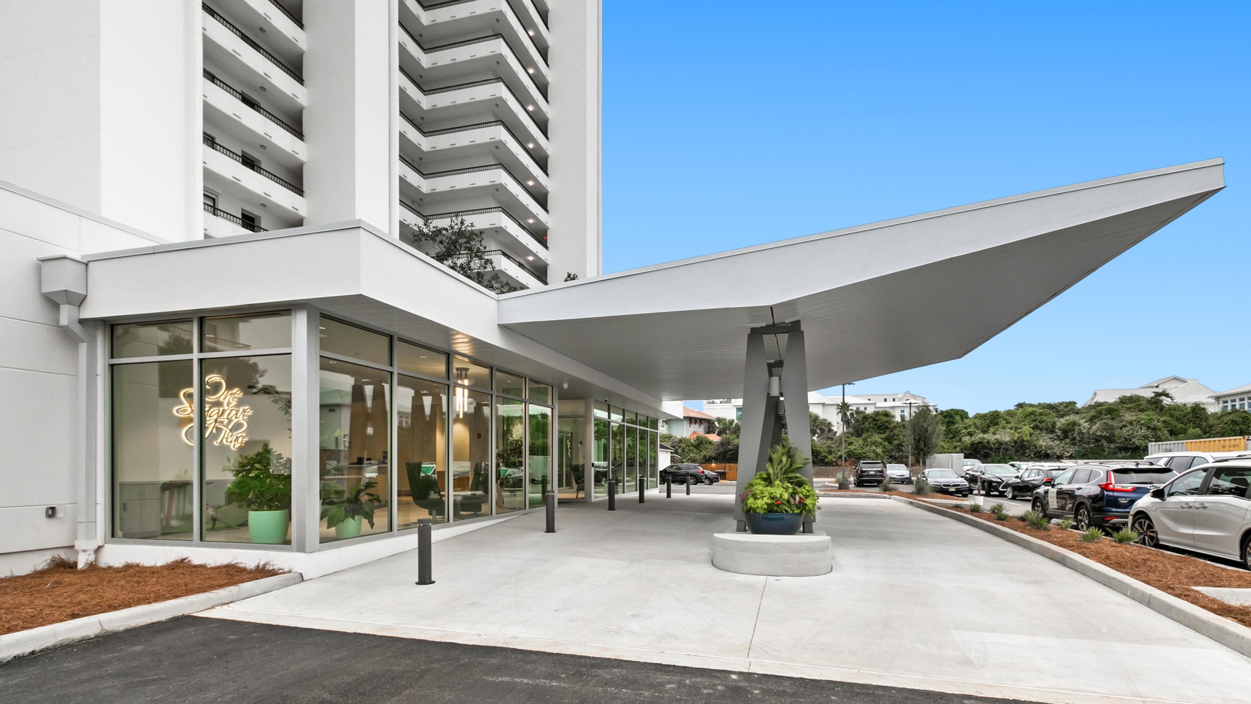 The modern entrance of a tall white condominium building features a striking angular canopy, a paved driveway, and a parking lot, surrounded by green landscaping.