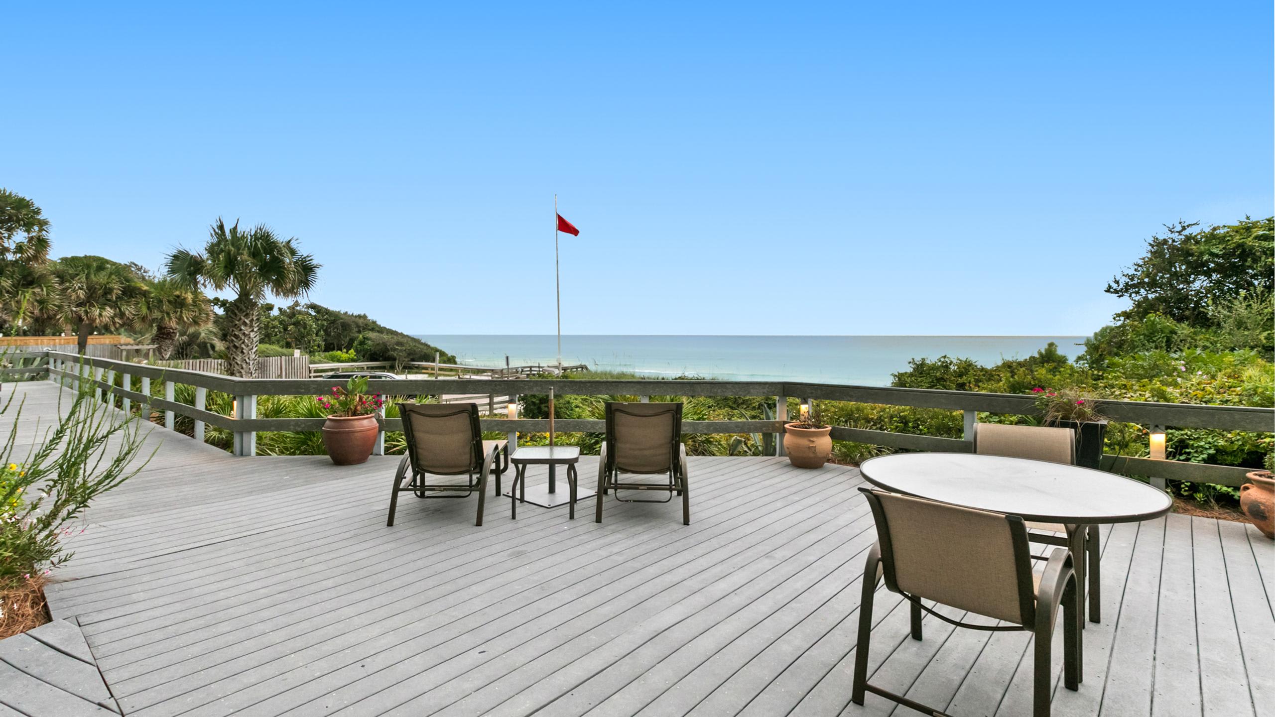 A spacious grey wooden deck furnished with lounge chairs and a dining set, offering expansive views of the calm blue ocean under a clear sky, with a red flag visible in the distance.