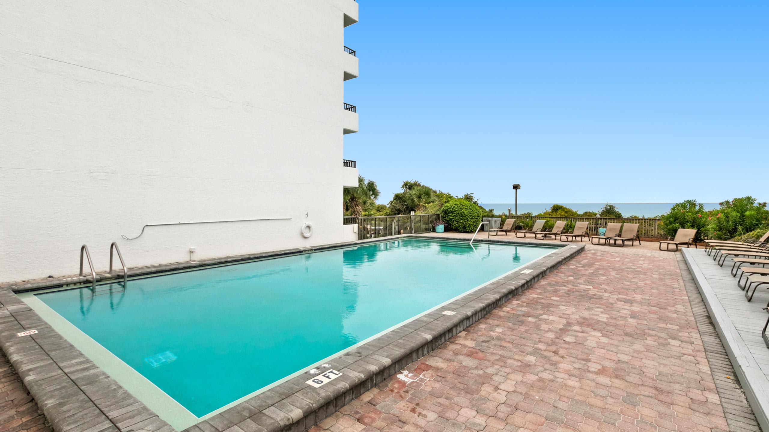 Rectangular outdoor swimming pool with a light blue interior, surrounded by a brick paver deck and numerous lounge chairs. A white building facade is visible to the left, and the ocean is in the distance.
