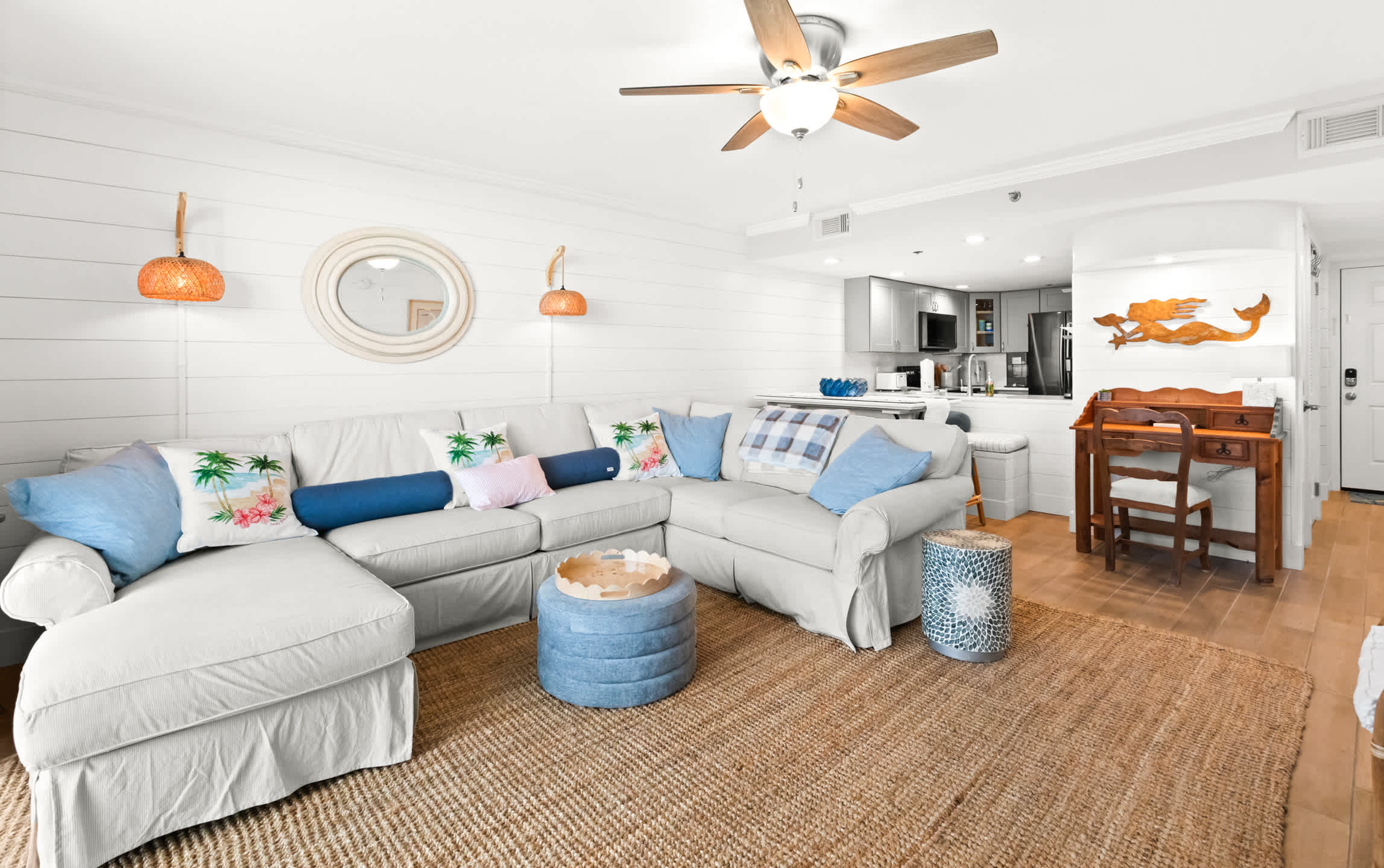Cozy living room with a large white sectional sofa, a natural jute rug, and shiplap walls, offering a view towards the open-concept kitchen and a small wooden desk area with a mermaid wall art.