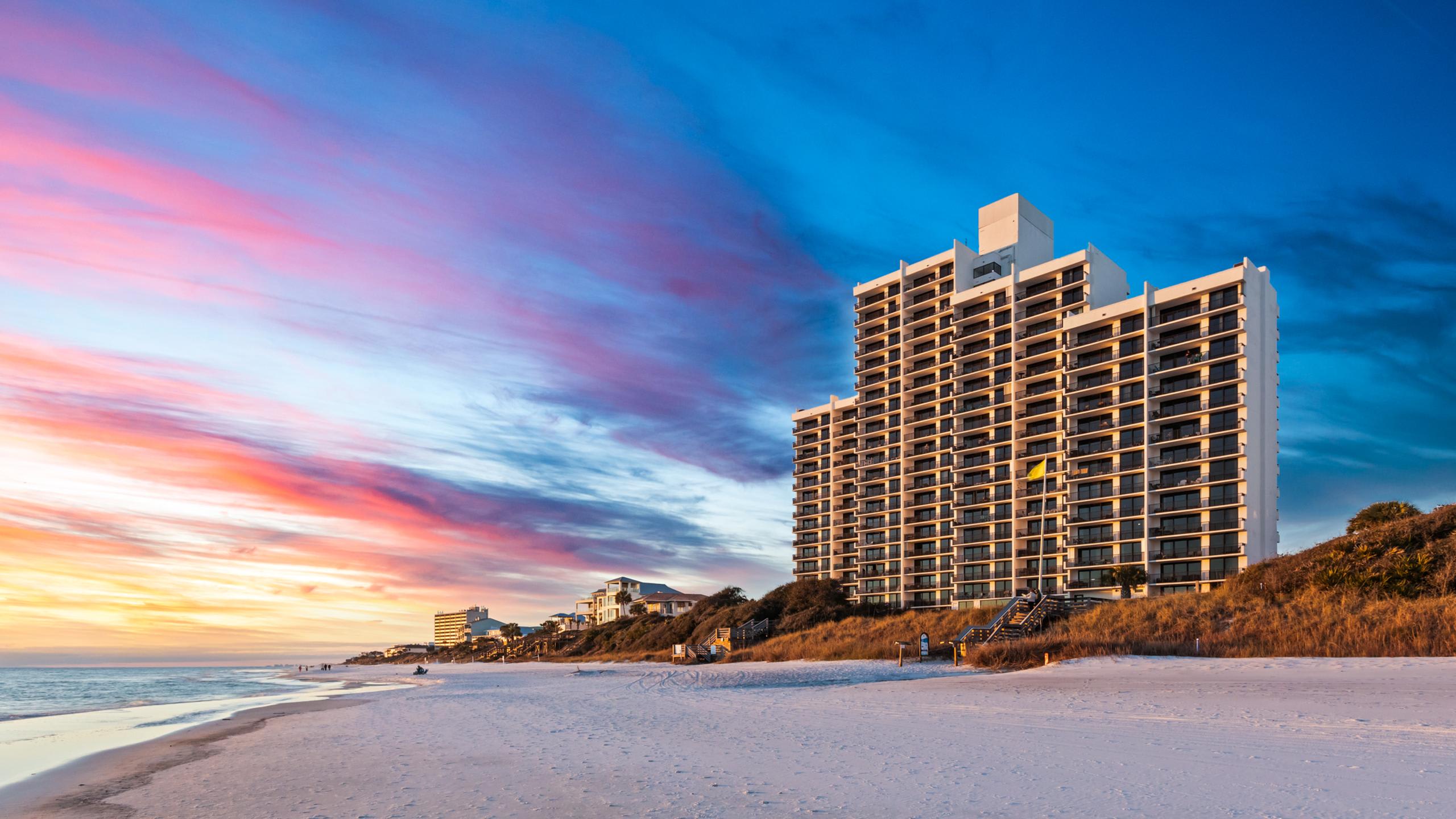 A large white condominium building with numerous balconies overlooks a wide sandy beach and the ocean, bathed in the warm glow of a colorful sunset sky.