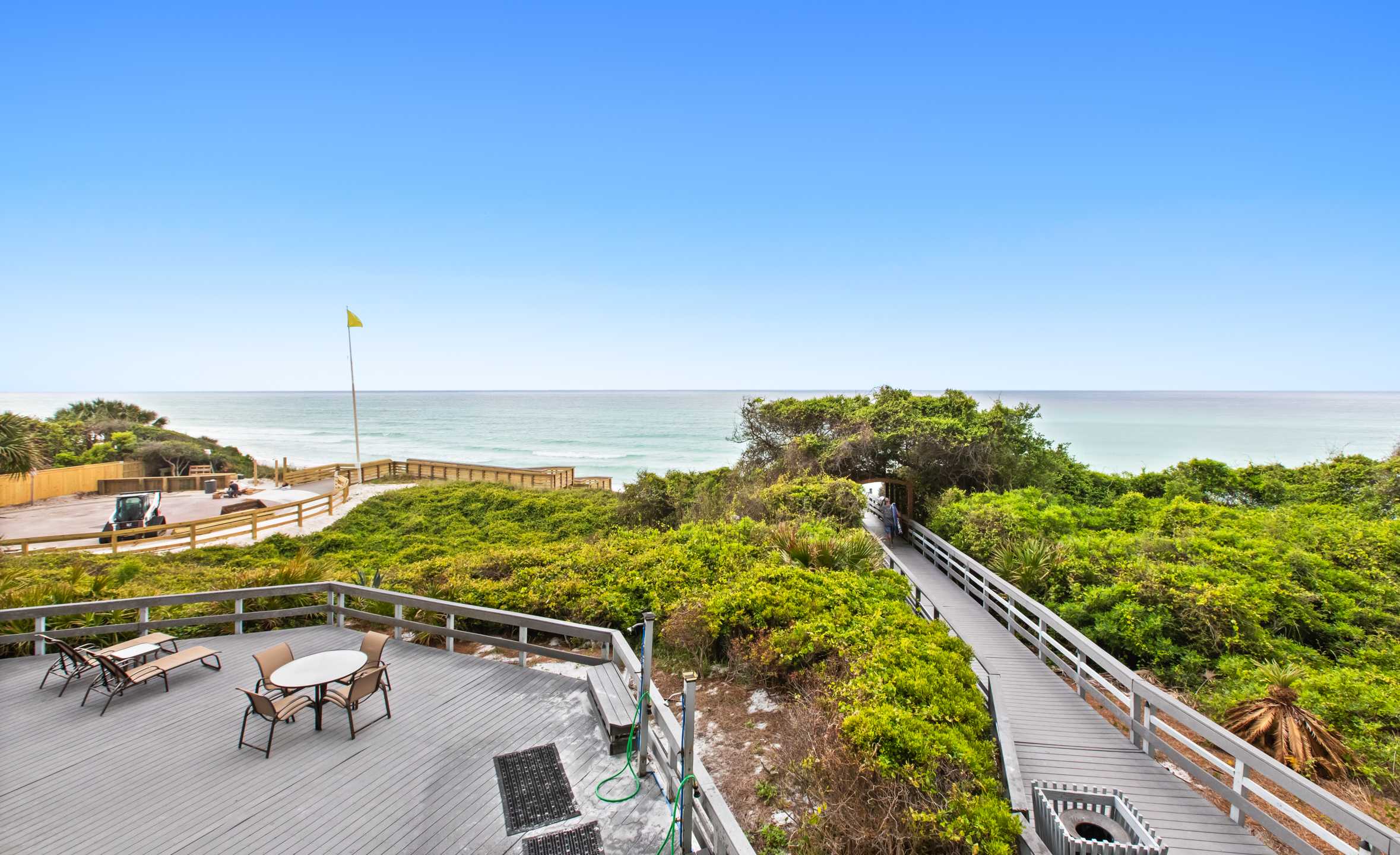 Expansive communal deck with light gray wooden planks and railings, offering panoramic views of the ocean and a sandy beach. Lush green vegetation surrounds a boardwalk leading to the beach, with a small outdoor dining set on the deck.