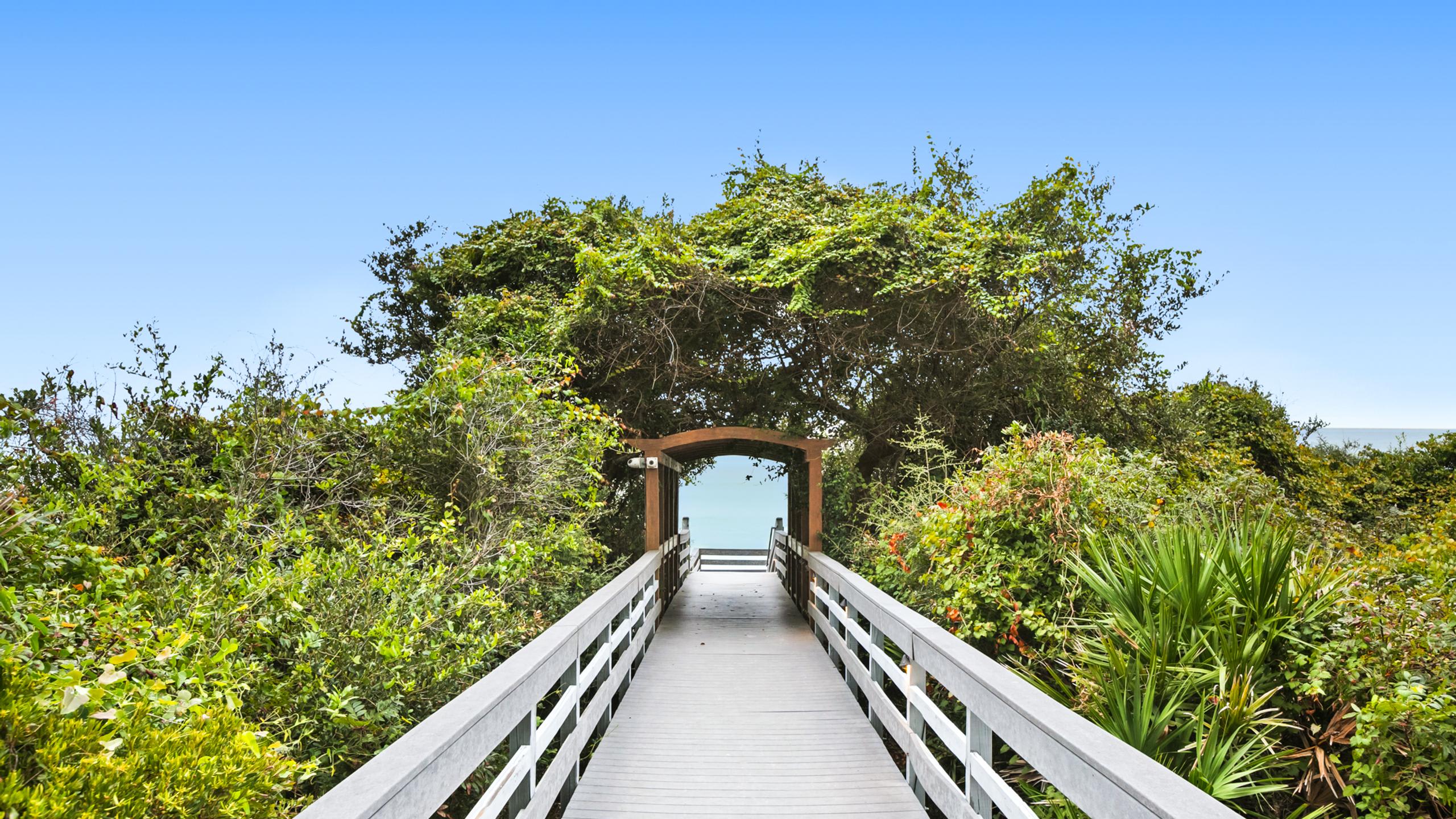 A wooden boardwalk with white railings extends through lush green foliage, leading towards an arched wooden gateway that frames a distant view of the blue ocean. The path is surrounded by dense, vibrant greenery and trees.