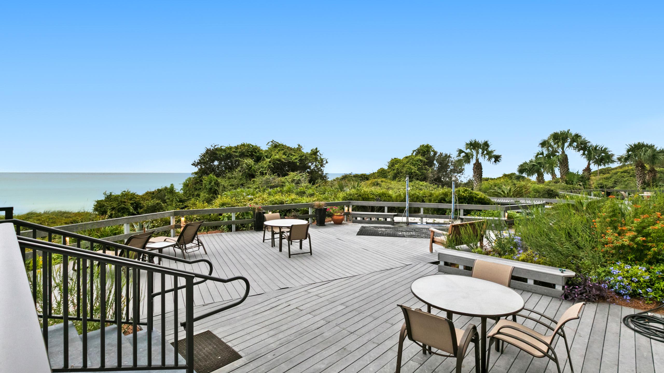 An expansive grey wooden deck features various outdoor dining and seating arrangements, surrounded by green foliage and offering a wide vista of the ocean horizon.