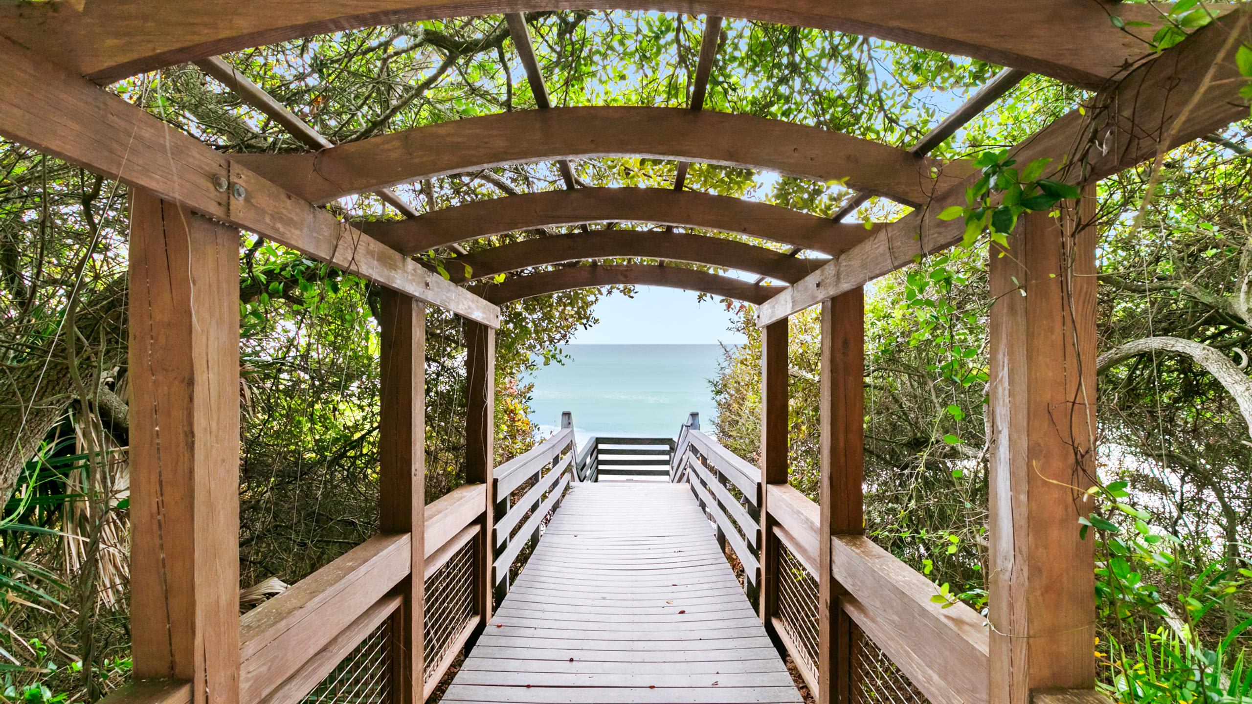 A rustic wooden boardwalk, covered by a leafy pergola, leads down towards the bright blue ocean, framed by dense green coastal vegetation on either side.