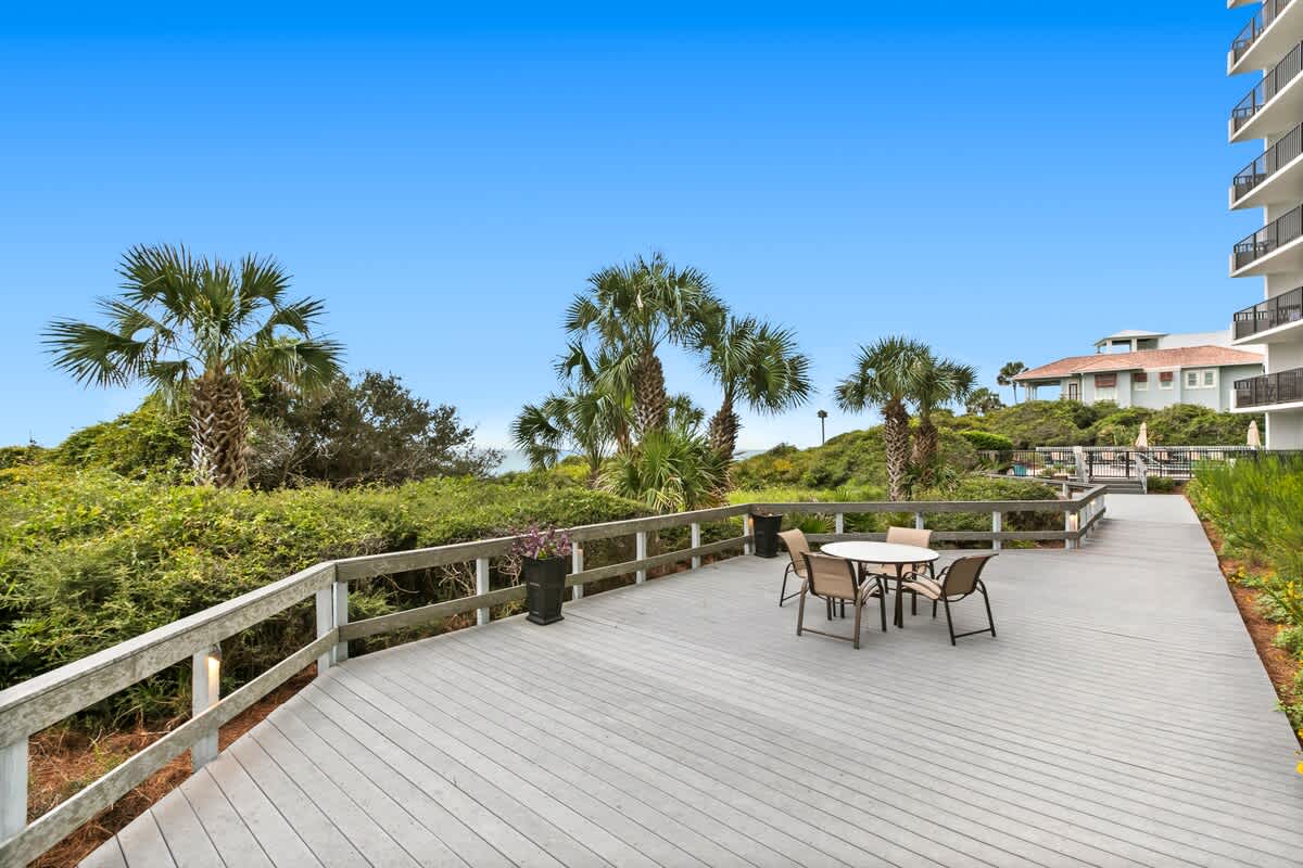 A wide gray composite deck with a white railing offers an outdoor dining set and potted plants, surrounded by vibrant green foliage and palm trees, with a tall white building visible in the background.