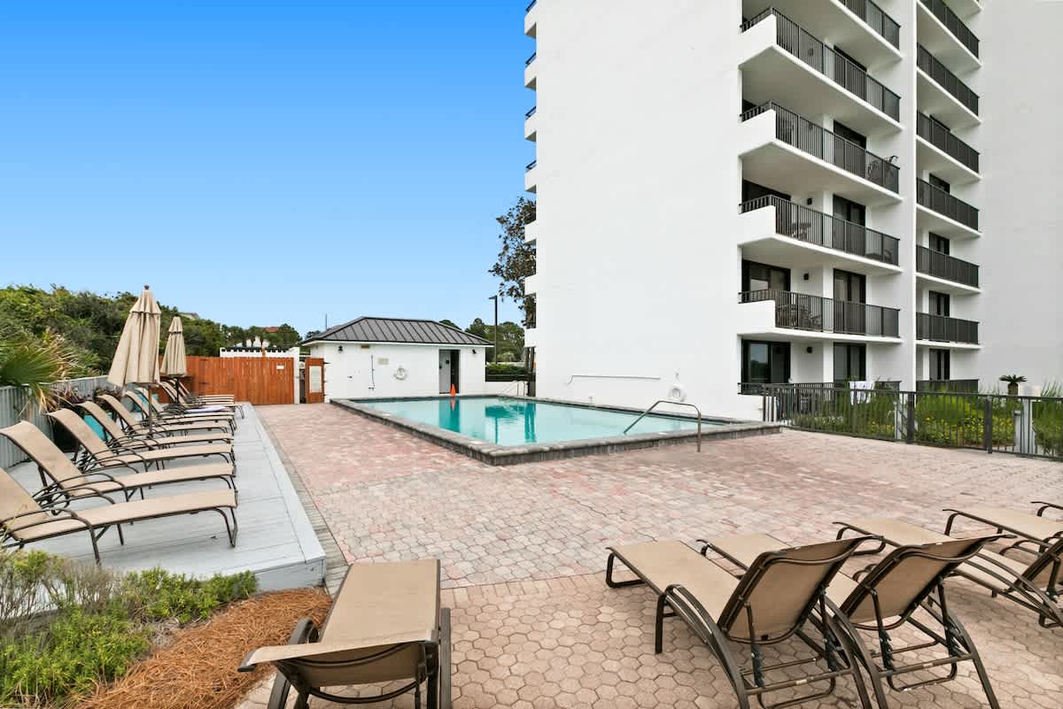 A rectangular outdoor swimming pool is centered within a spacious paver patio, lined with numerous lounge chairs and umbrellas, adjacent to a modern white high-rise building under a clear blue sky.