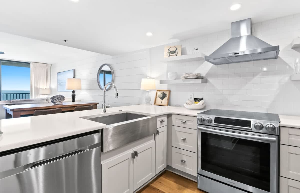 A modern kitchen boasts white subway tile backsplash, white shaker cabinets, stainless steel appliances including a range hood and oven, and a large farmhouse sink, with hardwood floors.