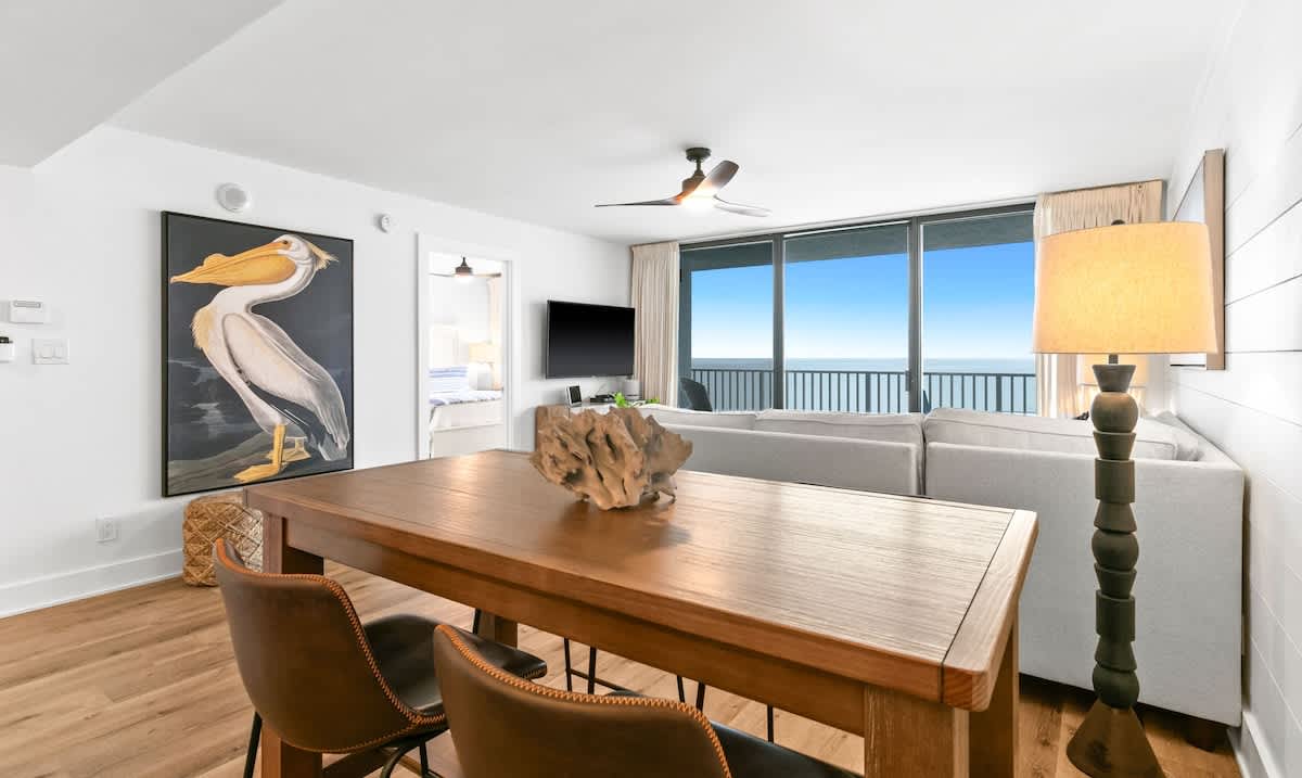 Spacious dining area with a rustic wooden table and brown leather chairs, featuring a large pelican artwork on the wall, and an open view to the living room and ocean beyond.
