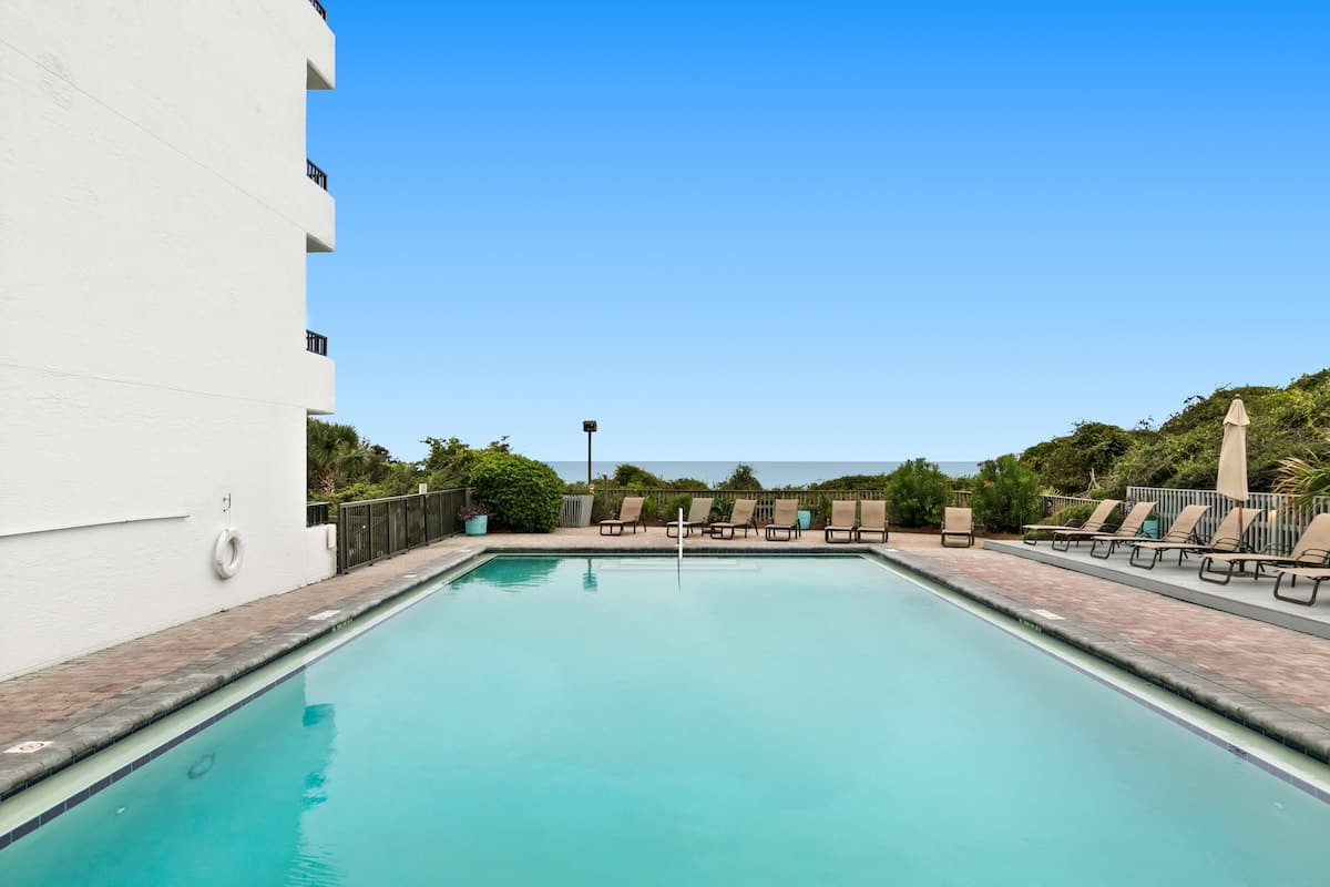 Outdoor swimming pool with blue water, surrounded by a brick paver deck featuring several lounge chairs, and a white building on the left, overlooking the ocean.
