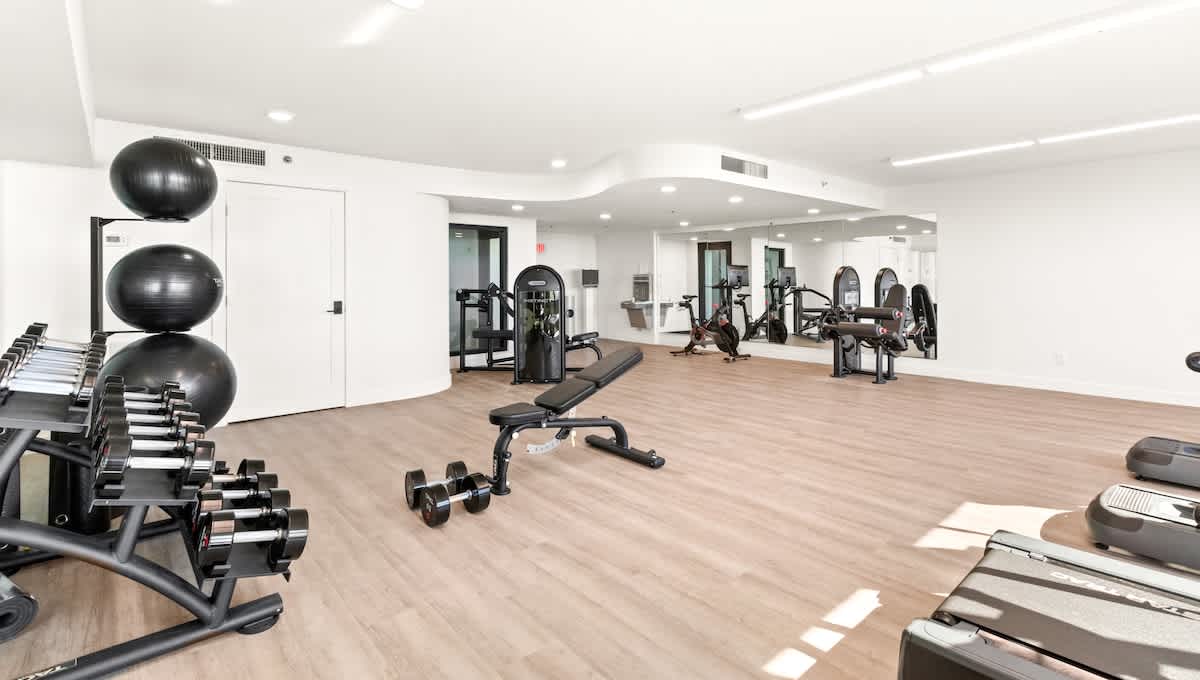 A spacious, well-lit fitness room with light wood-look flooring offers a variety of strength training equipment, including a dumbbell rack, weight bench, and resistance machines, with large mirrors reflecting the space.