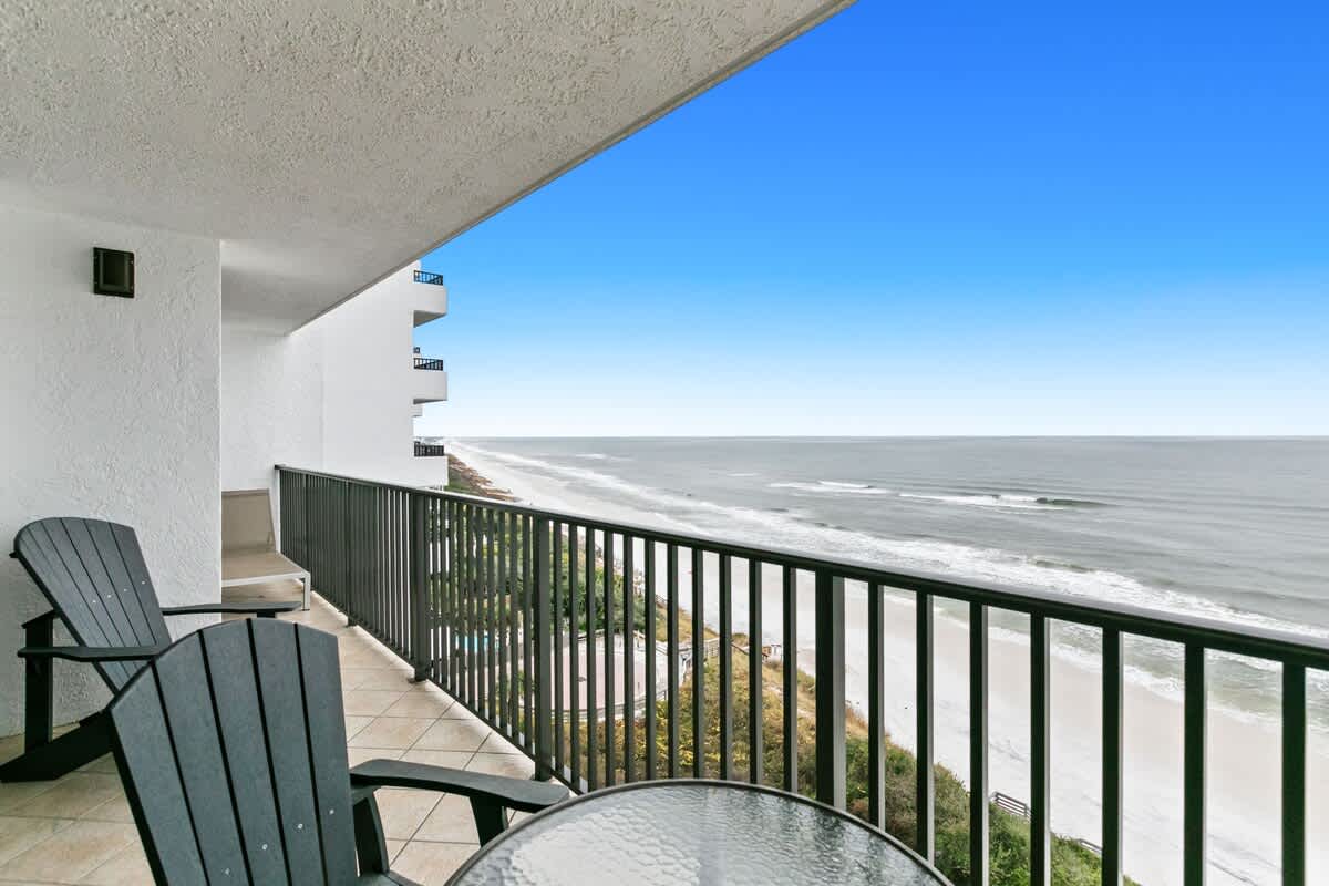This balcony view captures the extensive coastline with the ocean meeting the horizon and a white sandy beach below, featuring a glass-top table and two dark Adirondack chairs. The building's white exterior and multiple balconies are visible to the left.