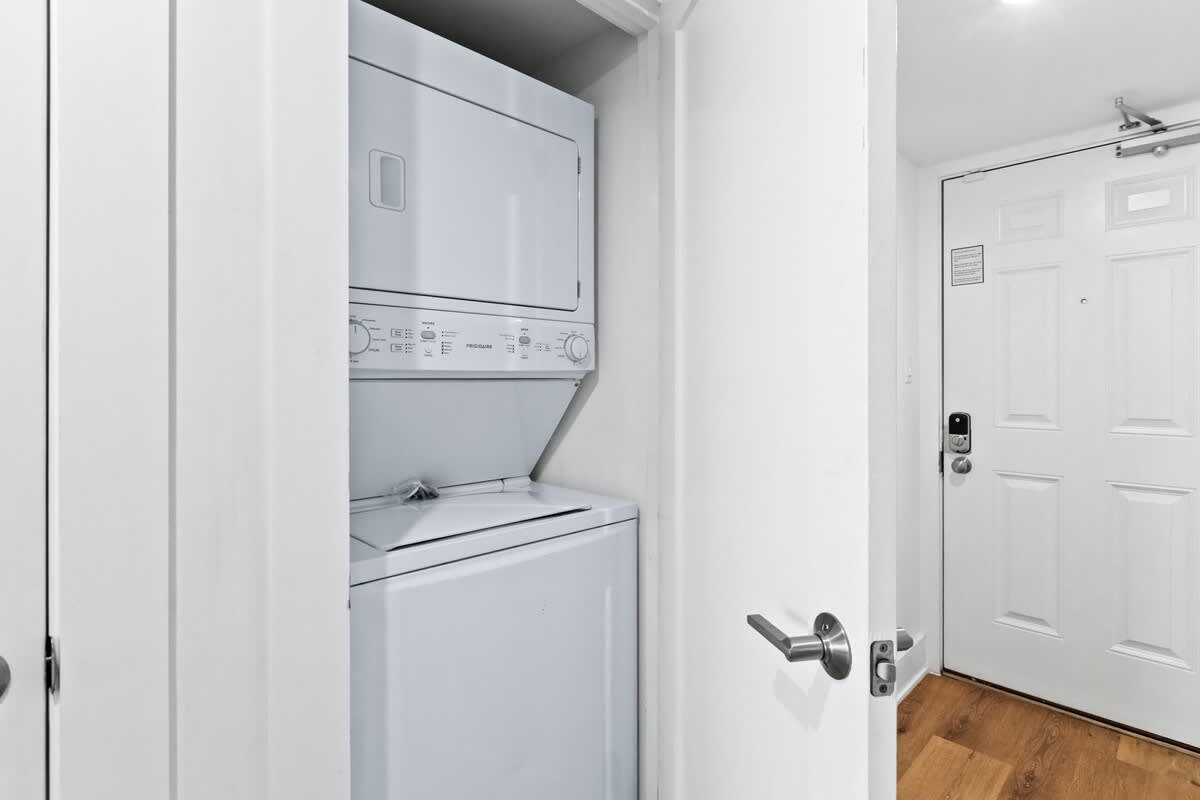 A compact laundry closet houses a white stacked washer and dryer unit, located next to a white entry door with a silver handle. The space features white walls and light wood-look flooring.